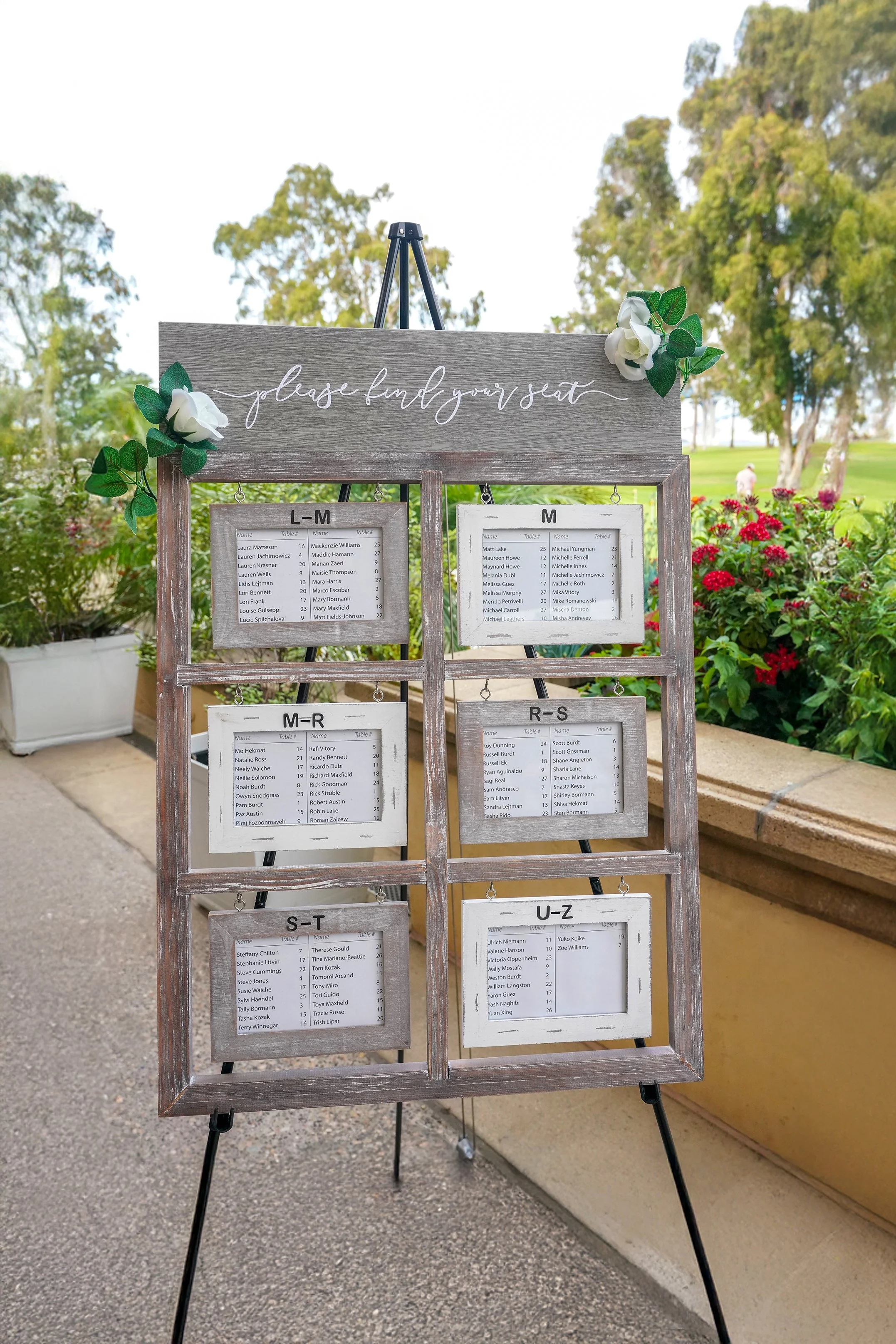 A wooden signboard with the words 'please find your seat' written on top, decorated with white flowers and green leaves. Below, several smaller placards display seating arrangements by alphabetical groups, set outdoors with trees and flowers in the b