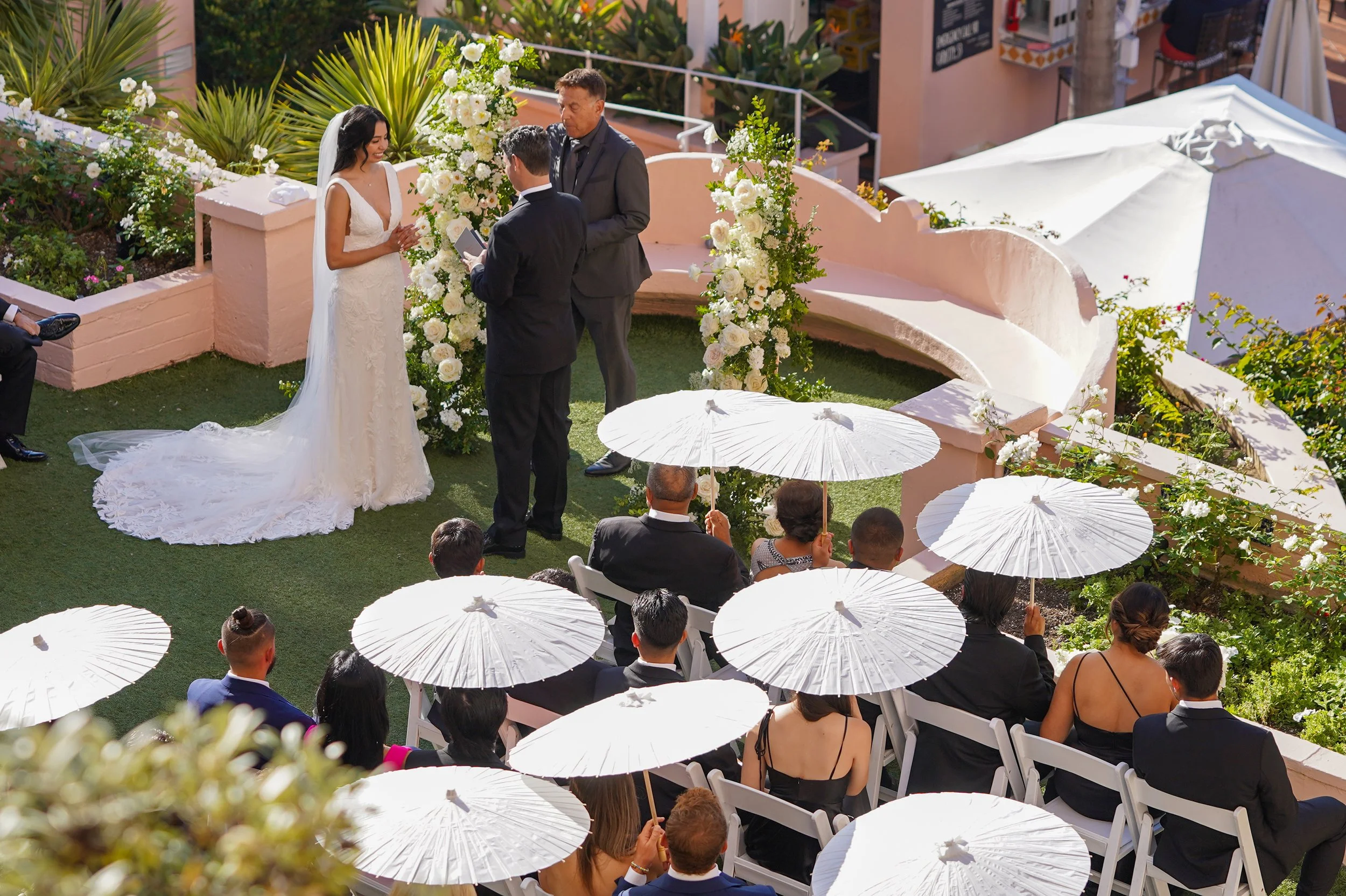 High-angle view of an outdoor wedding ceremony at La Valencia Hotel in La Jolla, showing wedding guests holding white paper parasol umbrellas while a bride and groom exchange vows in front of a white rose floral arch.