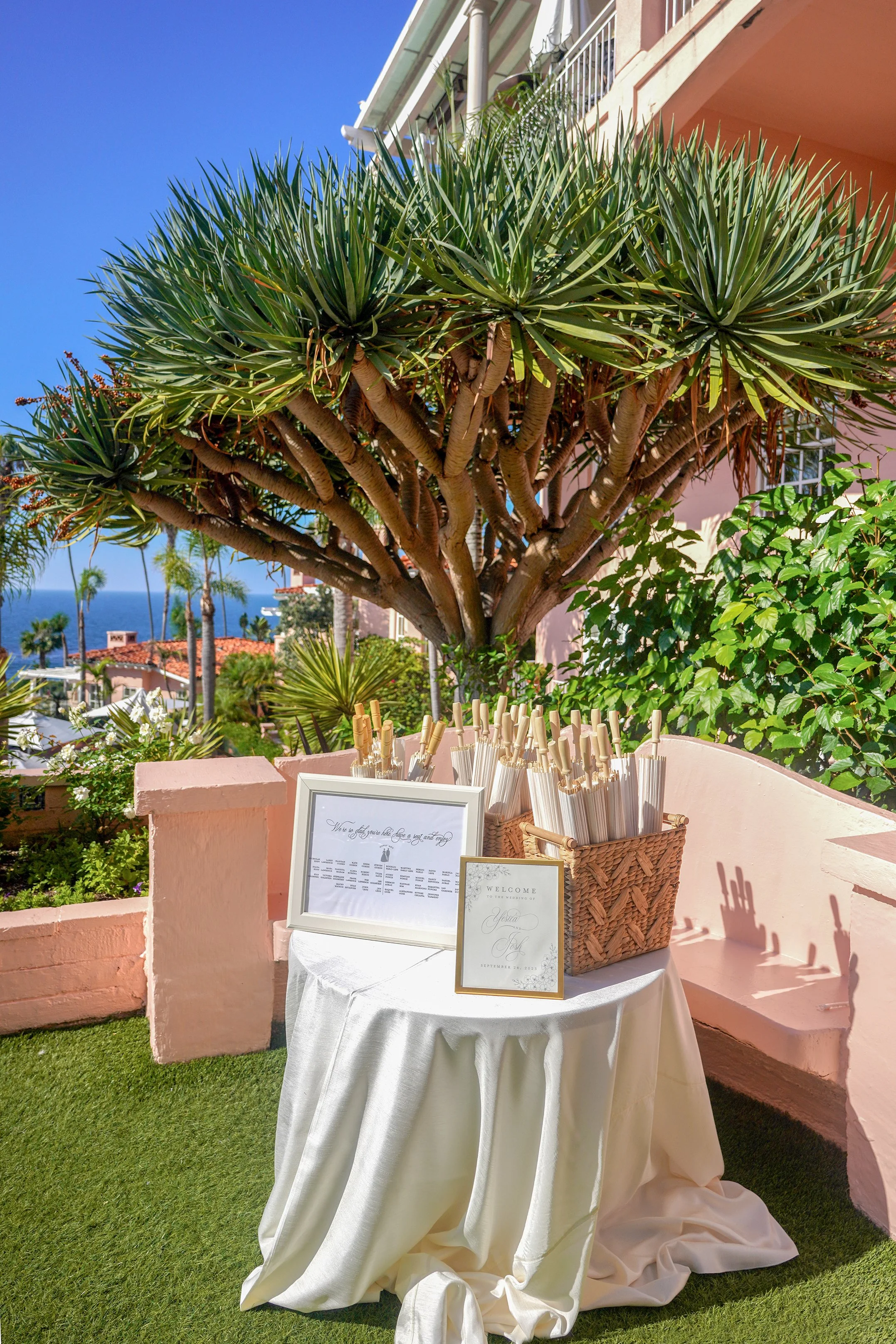 A white cloth-covered welcome table is set up outdoors at La Valencia Hotel under a large, unique dragon tree in La Jolla, California.