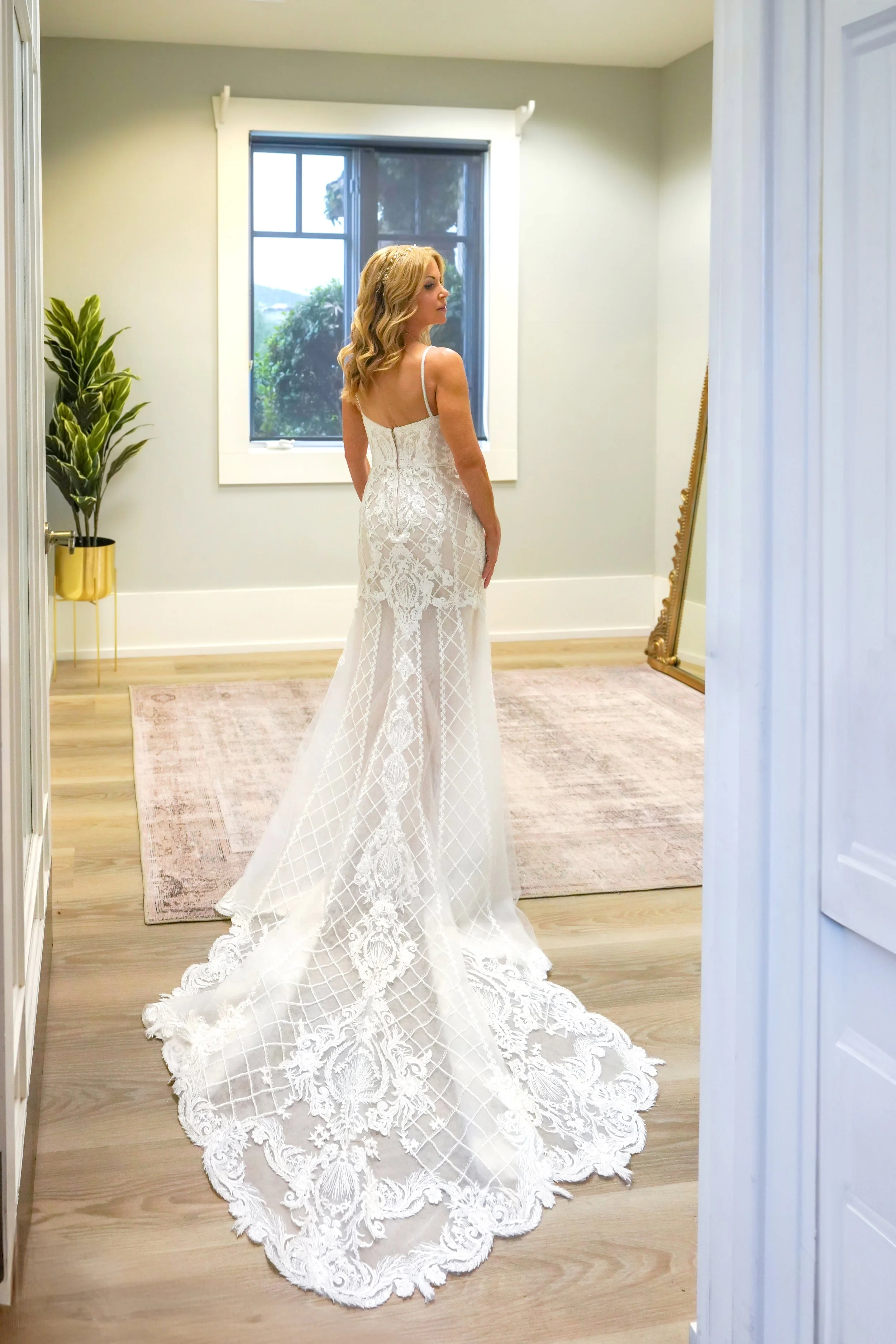 Full-length back view of a blonde bride modeling a fitted lace wedding dress with an intricate geometric and floral train.