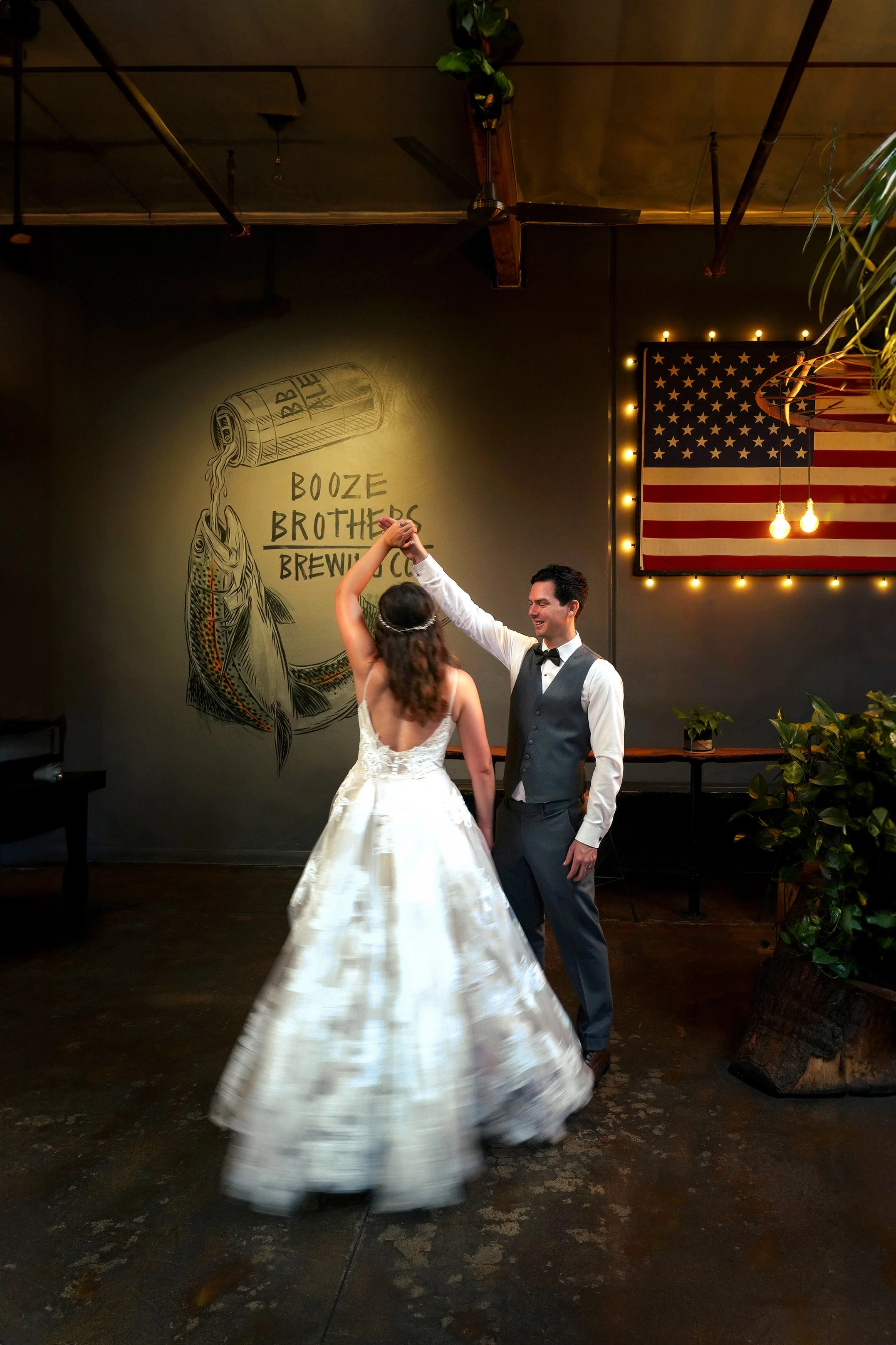 The bride and groom shares their first dance inside Booze Brothers Brewing Co. The bride, seems from behind in a flowing white, low-back wedding gown, twirls as the groom, dressed in a gray vest, trousers, and bow tie, smiles and hold her hand high.