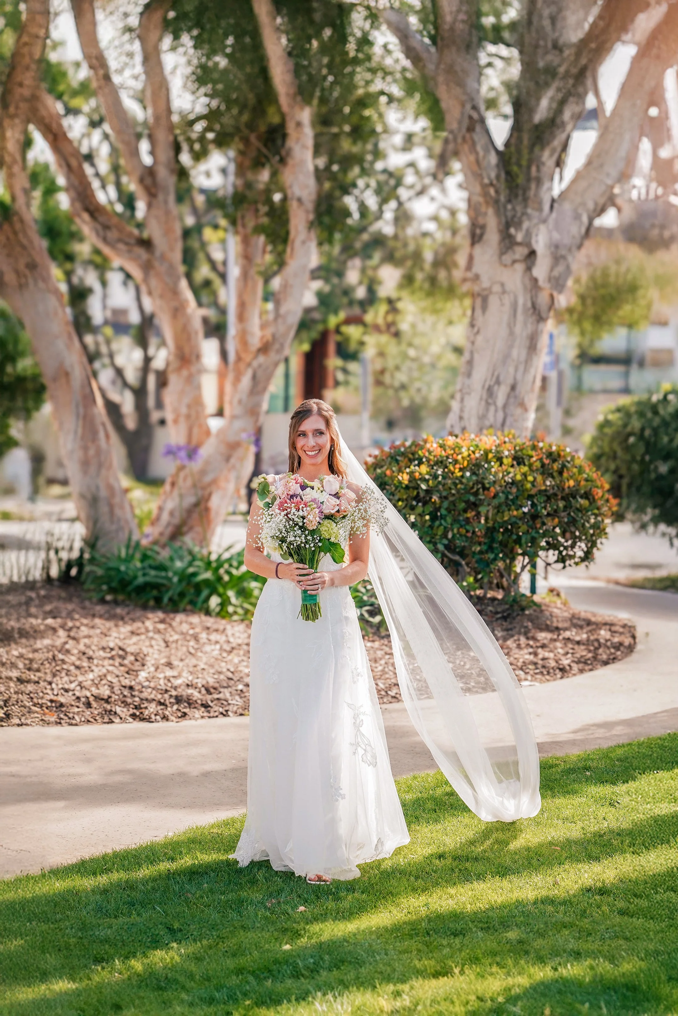 A smiling bride in a white wedding dress and veil holding a bouquet of pink, purple, and white flowers stands outside in a garden, with large trees and green shrubbery in the background.