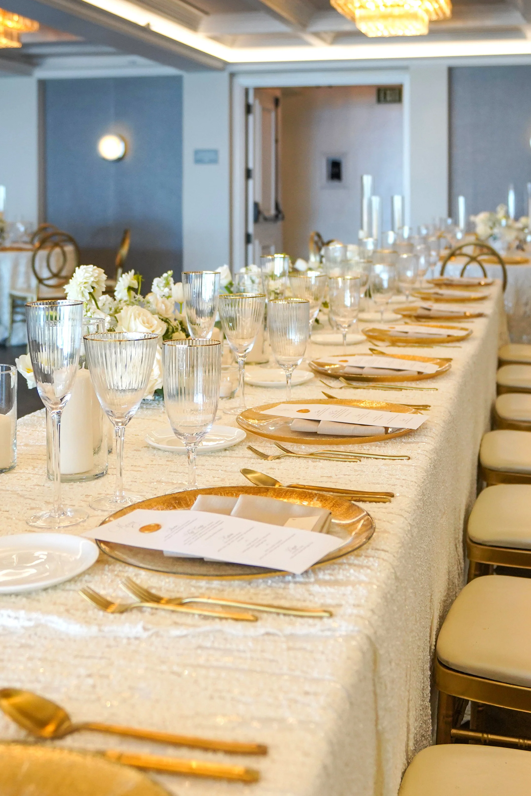 A long banquet table set up for a luxury indoor wedding reception. The table features shimmering white sequin tablecloths, custom white menu cards with gold wax seals, textured metallic gold charger plates, and matching gold flatware. Ribbed crystal 
