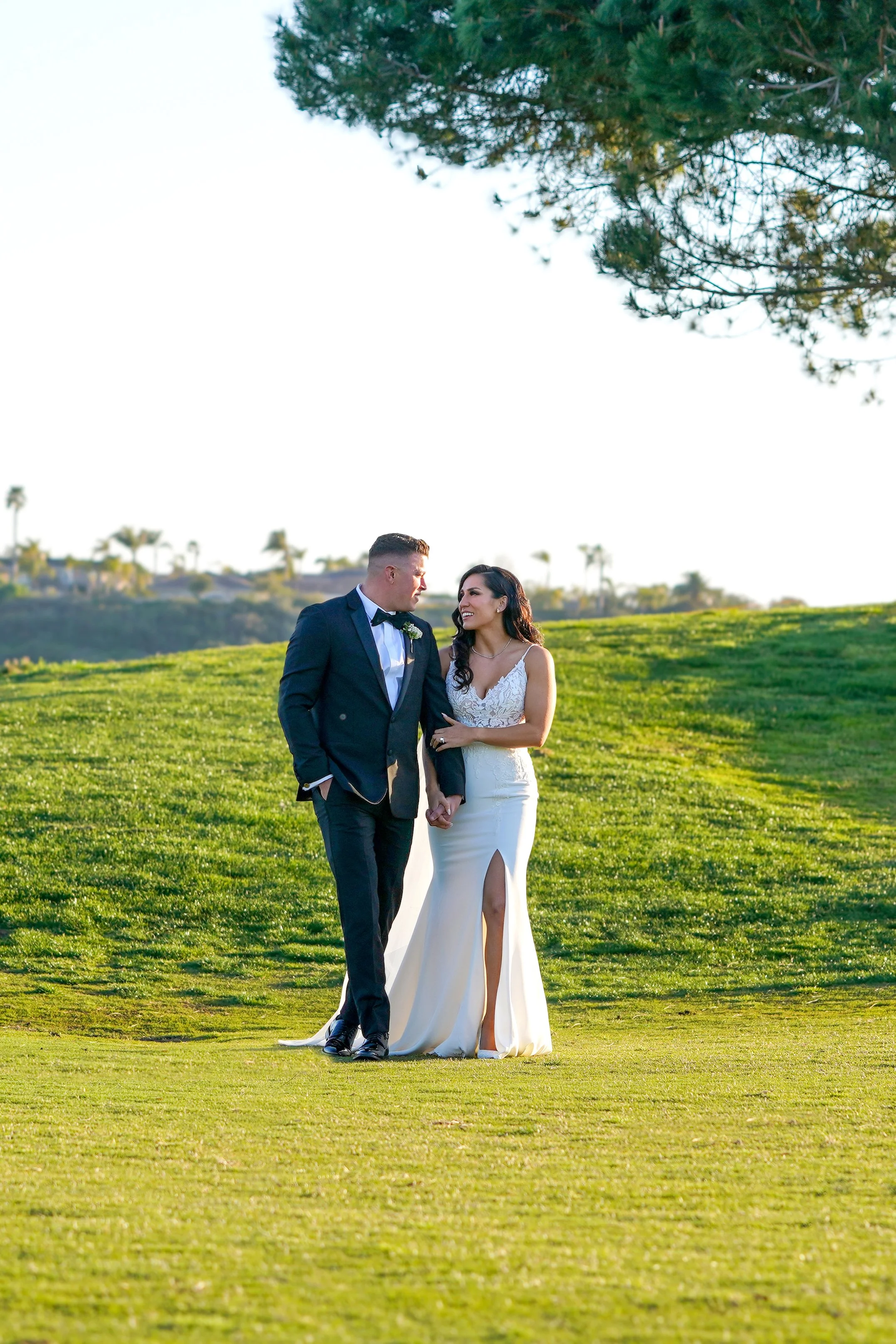 Bride in a lace bodice dress with a high slit and groom in a black tuxedo holding hands while walking on a grassy golf course.
