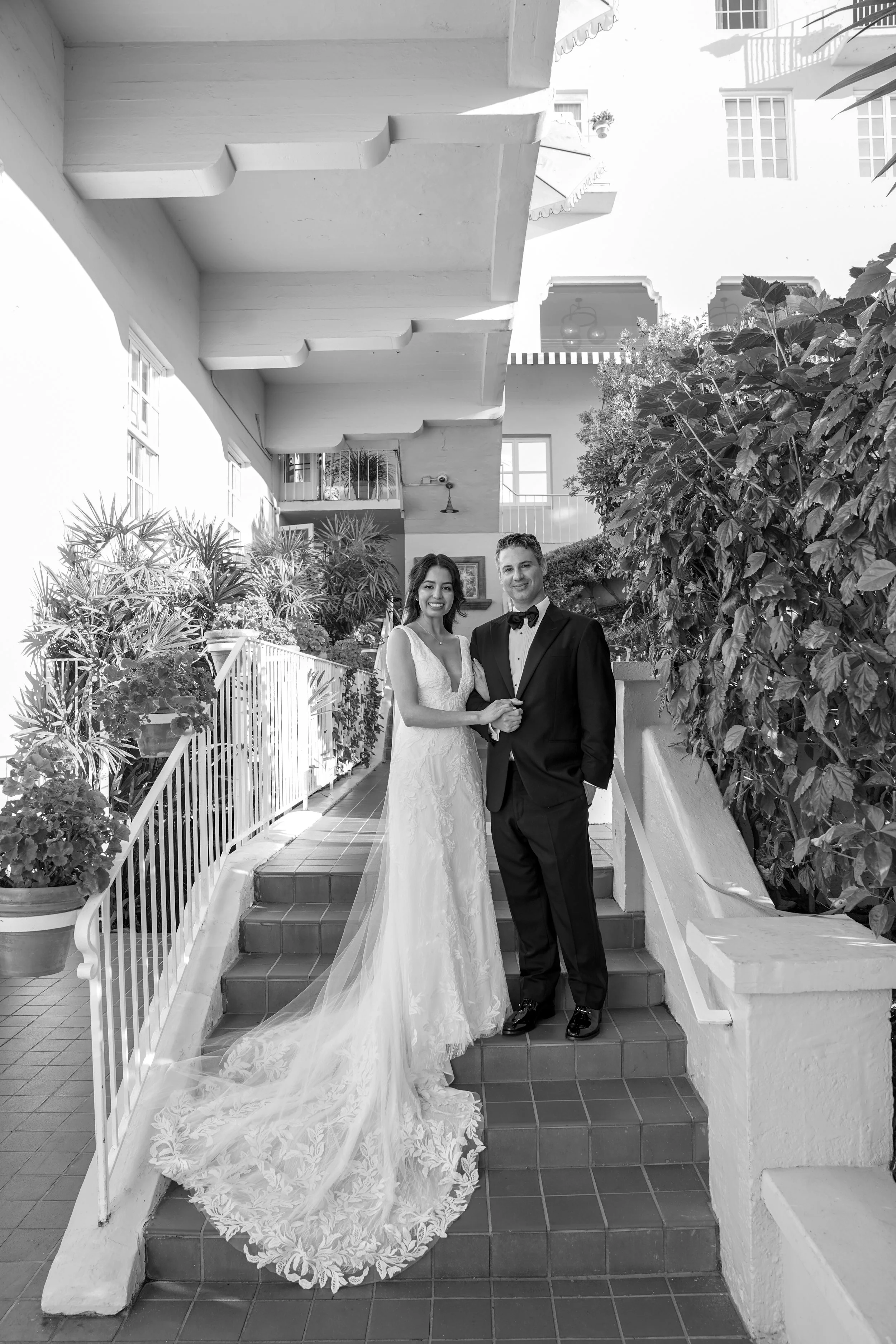 A smiling bride wearing an A-line lace wedding gown with a dramatic cathedral-length lace train alongside her groom.