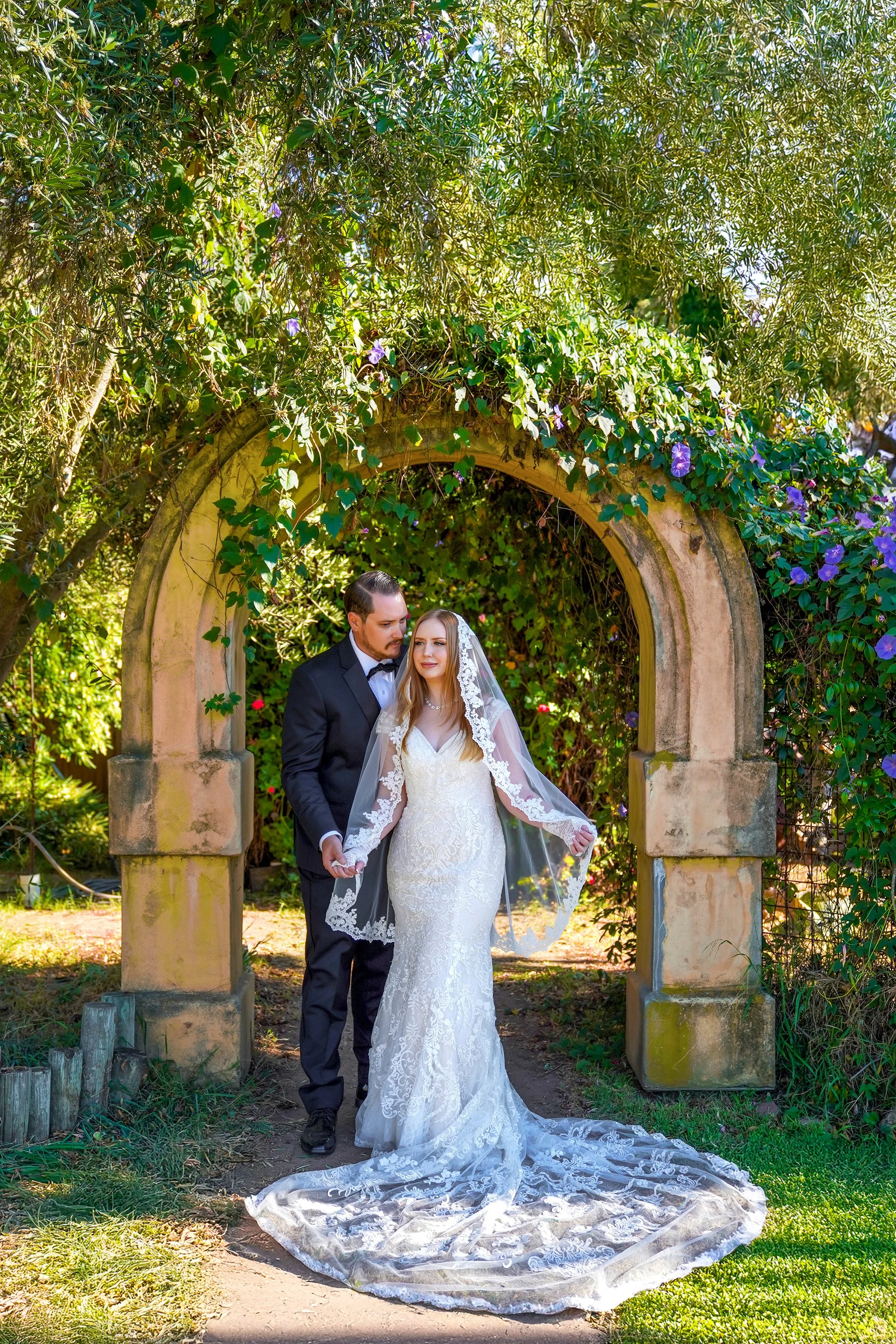 Outdoor wedding portrait of a bride in a lace mermaid wedding dress and long lace veil with a groom in a black tuxedo, posing under an ivy-covered stone archway.