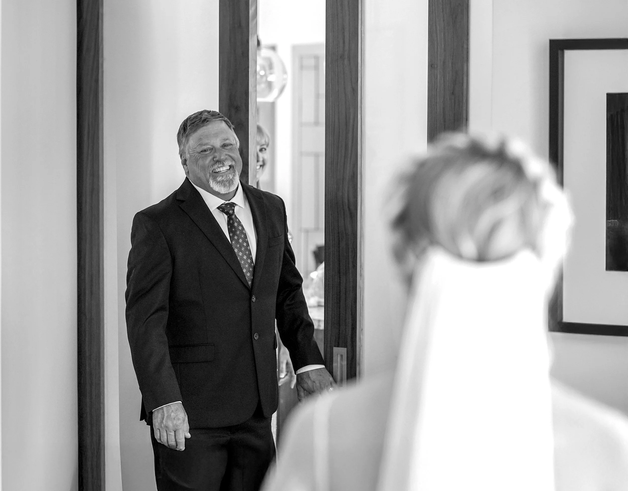 Father's joy during wedding day first look. Standing in the doorway, he beams with pride and emotion as he sees his daughter as a bride for the first time. The bride's veil and gown are softly blurred in the foreground.
