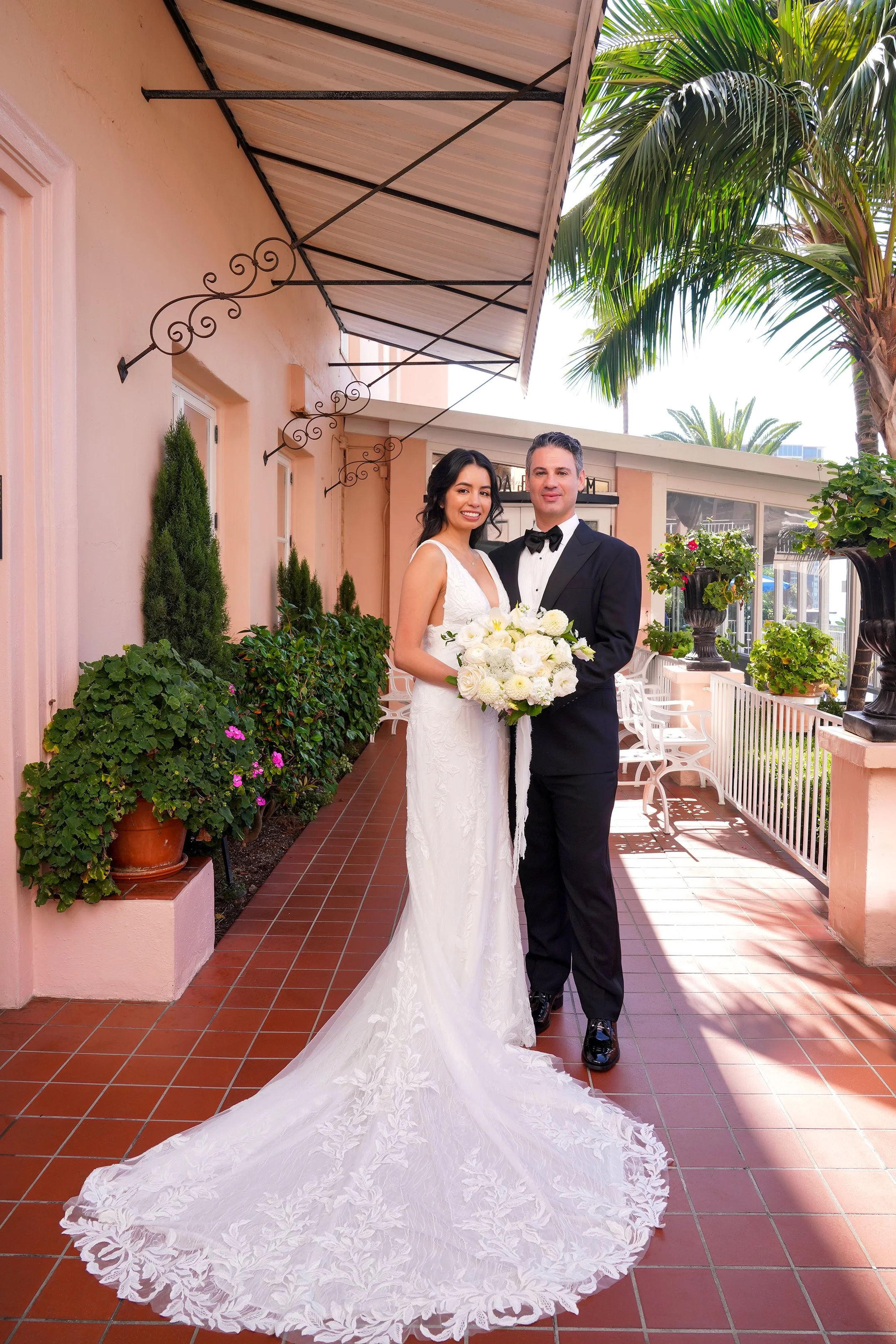Bride in a V-neck lace wedding dress and groom in a black tuxedo holding a white rose bouquet outside the Veranda Ballroom at La Valencia Hotel in La Jolla, San Diego.