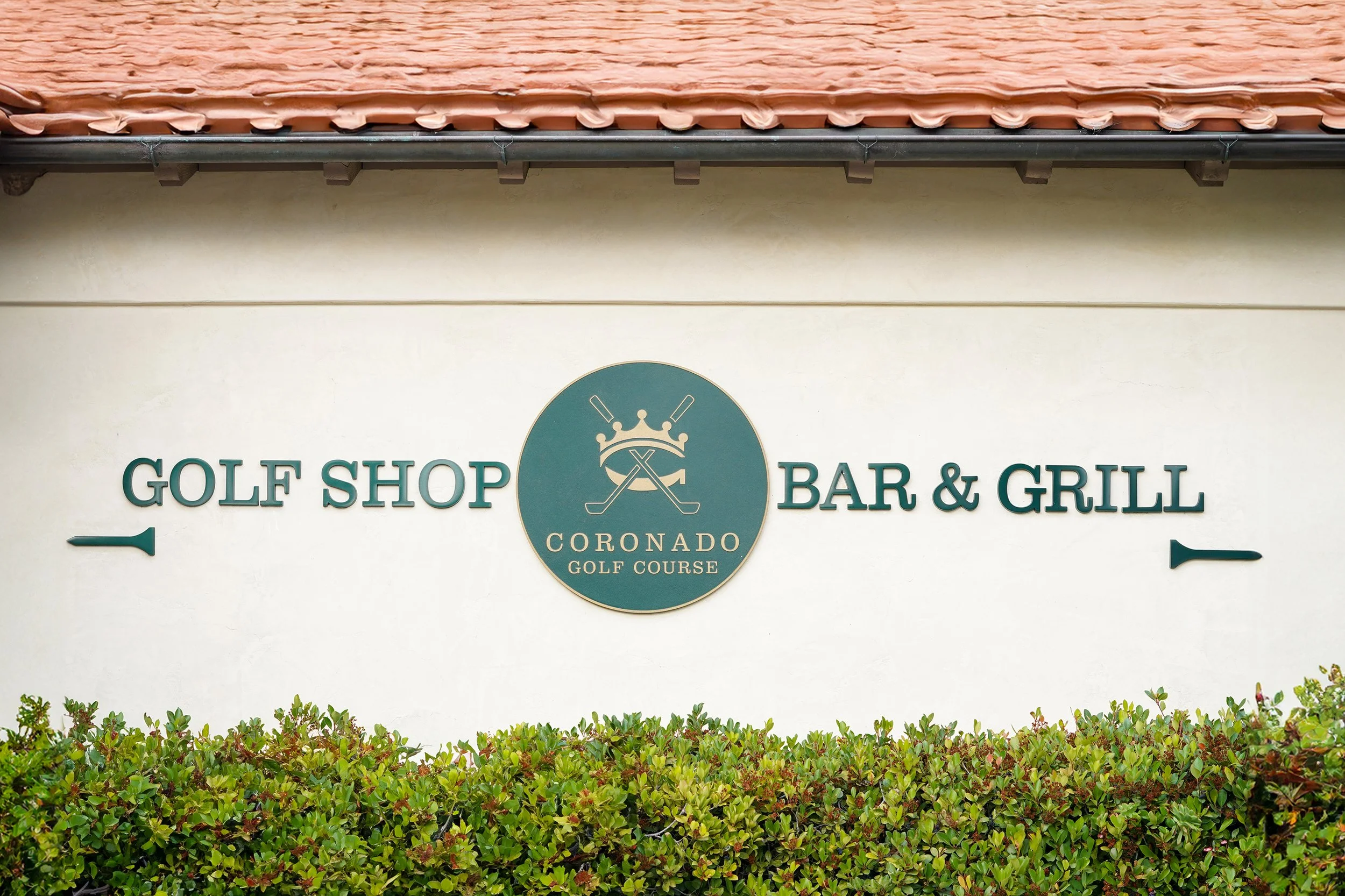 Sign for a golf shop, bar, and grill at the Coronado Golf Course with greenery in front.