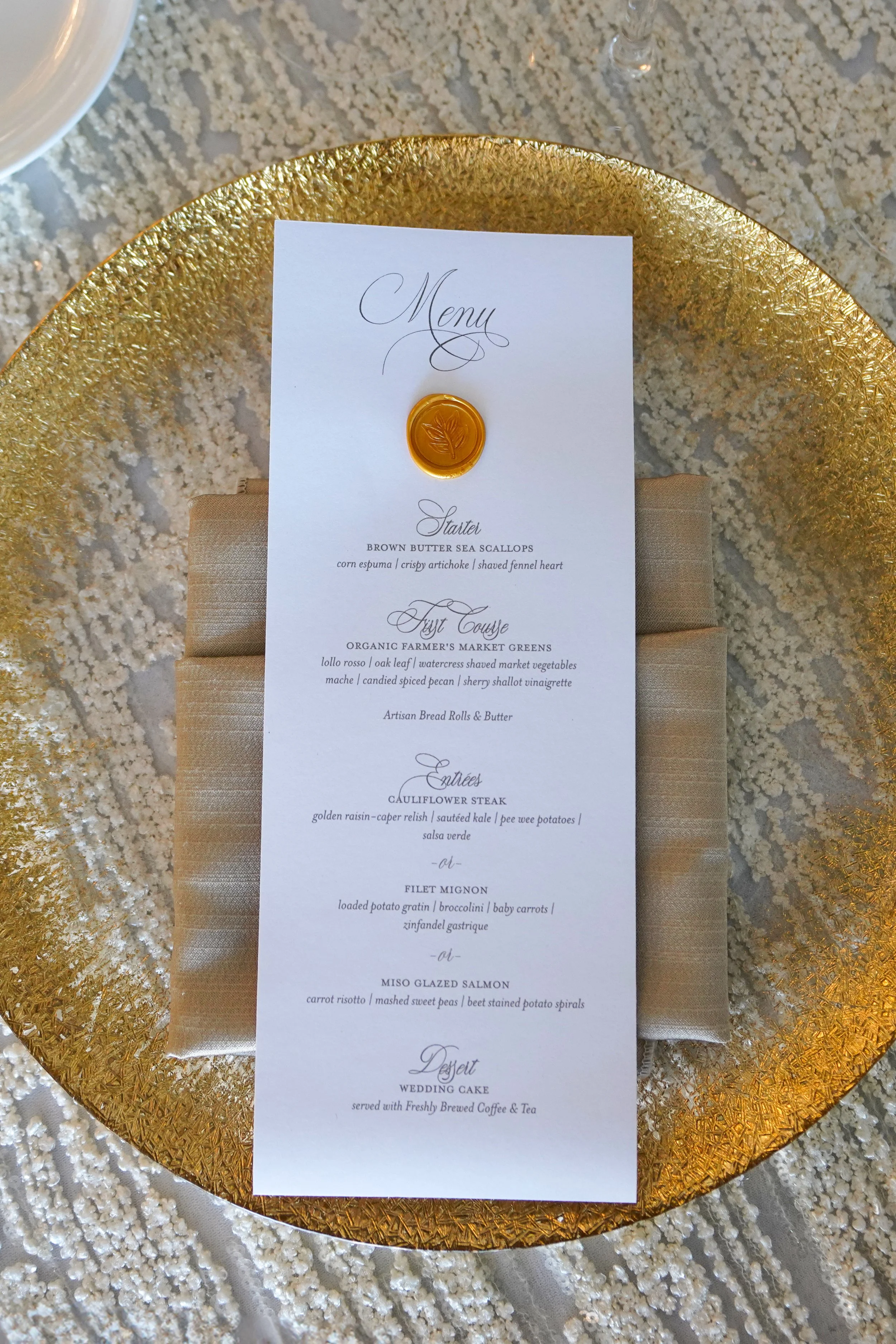 A top-down view of an elegant wedding reception table setting detail. A tall, white custom wedding menu card features sophisticated script typography, a gold leaf wax seal stamp, and a multi-course dinner menu including brown butter sea scallops, fil