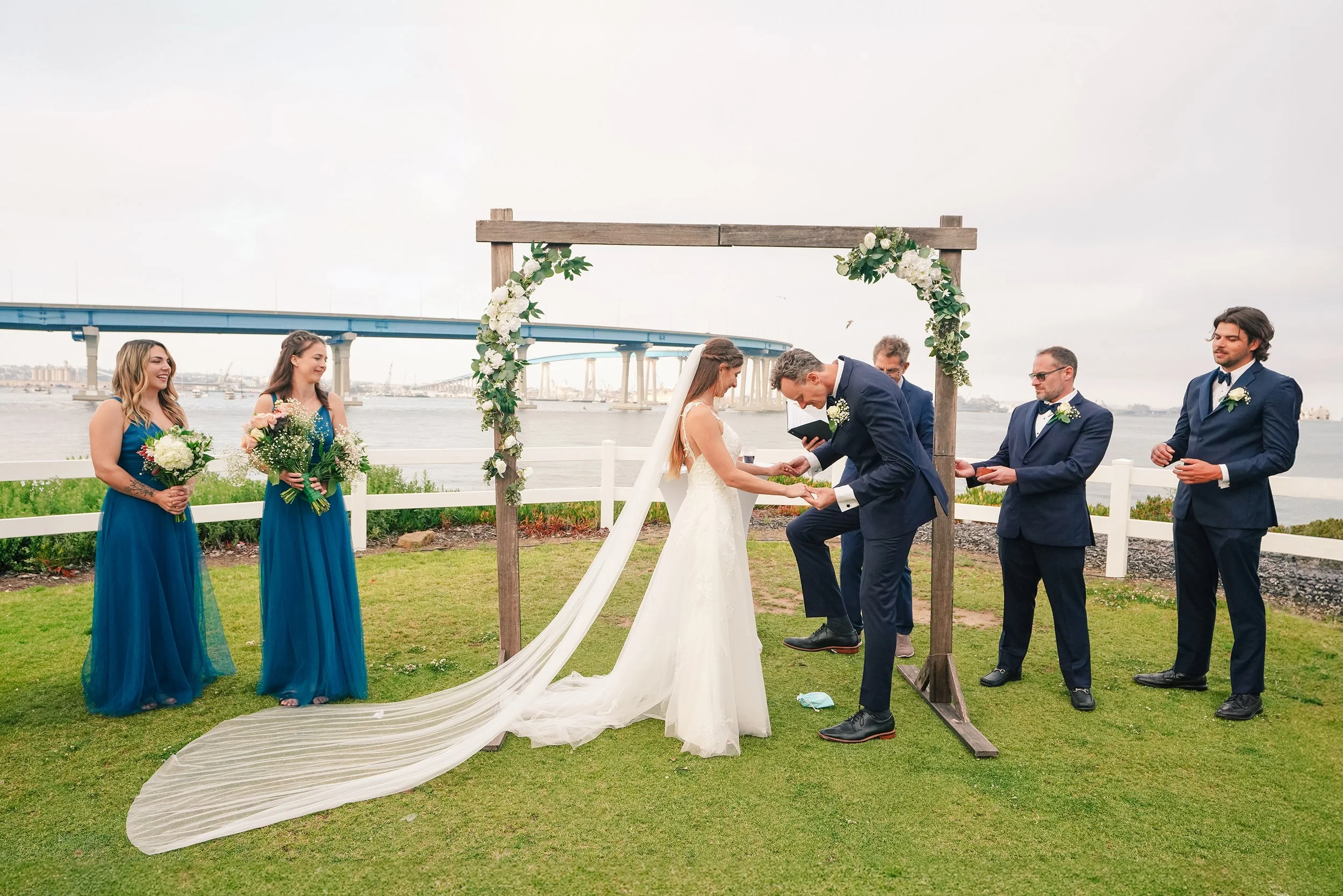 A wedding ceremony outdoors by the water with a bridge in the background. The bride and groom are exchanging rings under a wooden arbor decorated with white flowers and green foliage, while three groomsmen and two bridesmaids stand nearby.