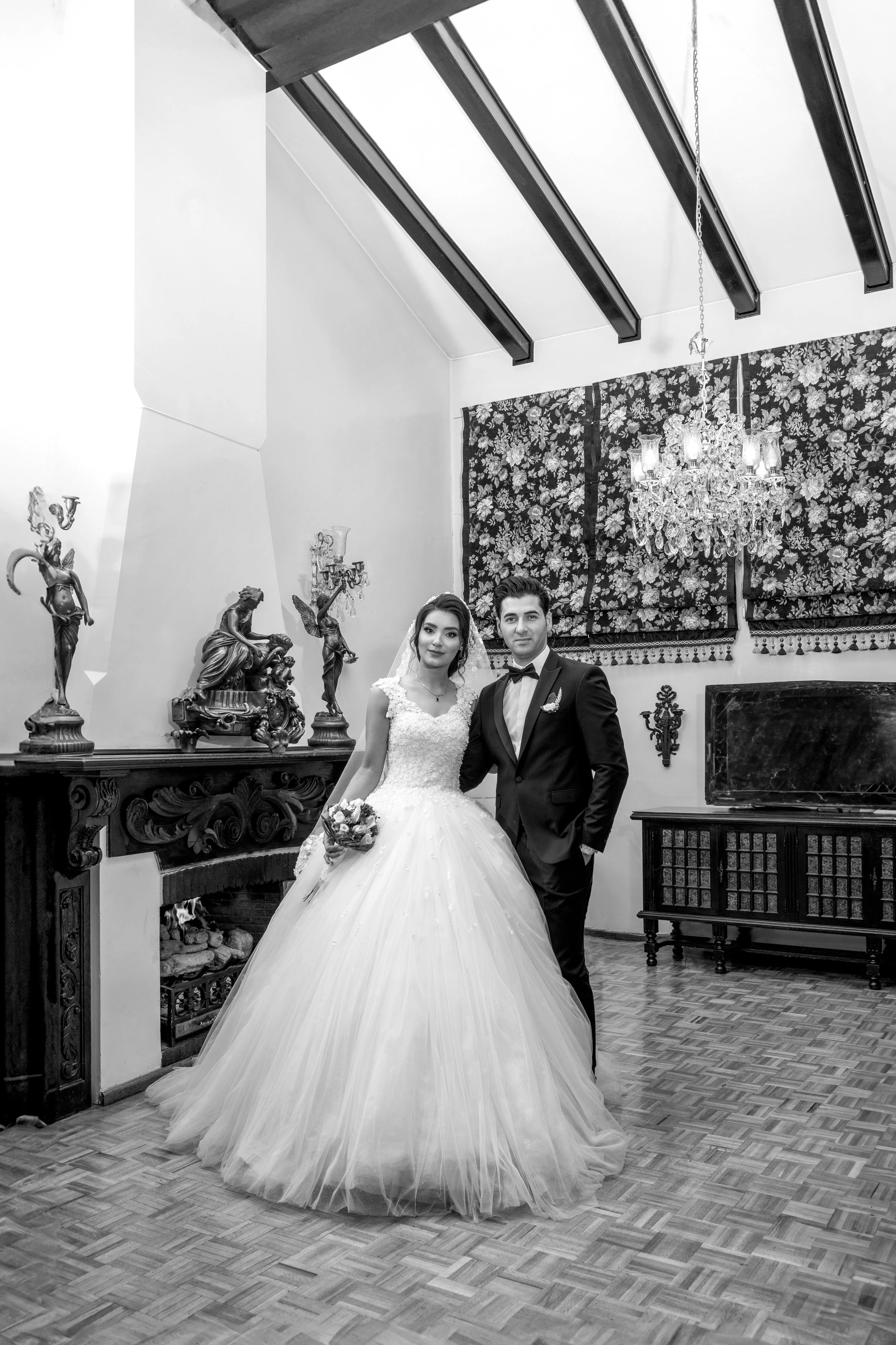 Classic black and white wedding portrait of a bride in a tulle ballgown and a groom in a tuxedo inside a luxury mansion venue with an ornate fireplace and crystal chandelier.