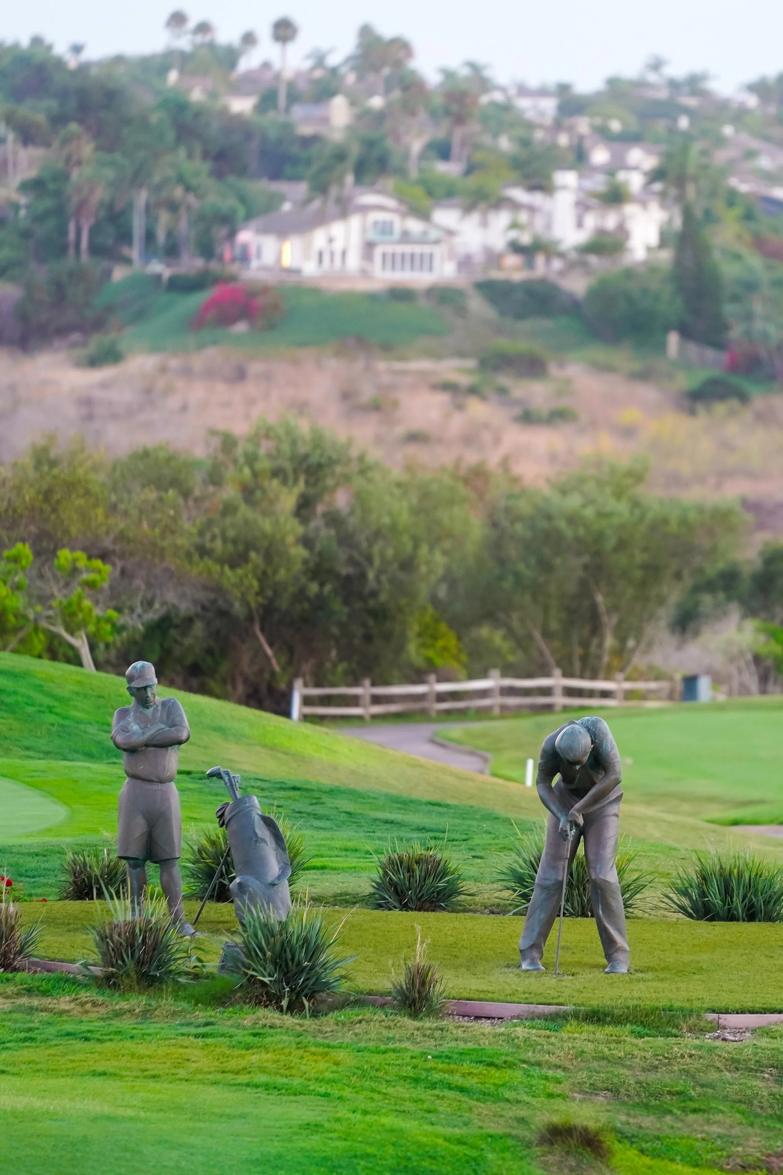 Lifelike bronze sculptures of a golfer putting and a caddie watching on a green fairway at The Crossings at Carlsbad with coastal hillside homes in the background.
