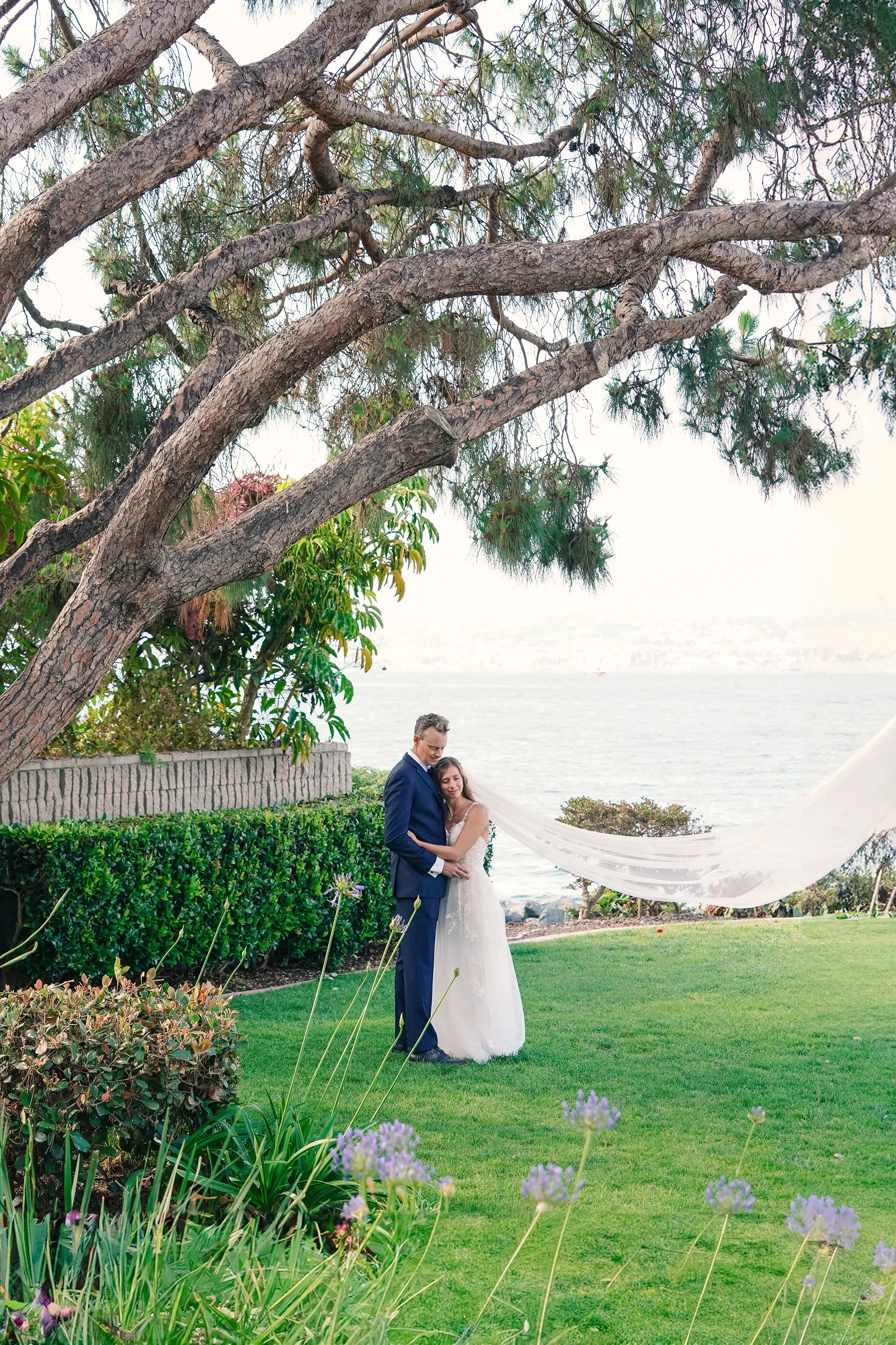 A bride and groom standing together outdoors near a body of water, surrounded by greenery and flowers, with large tree branches overhead.