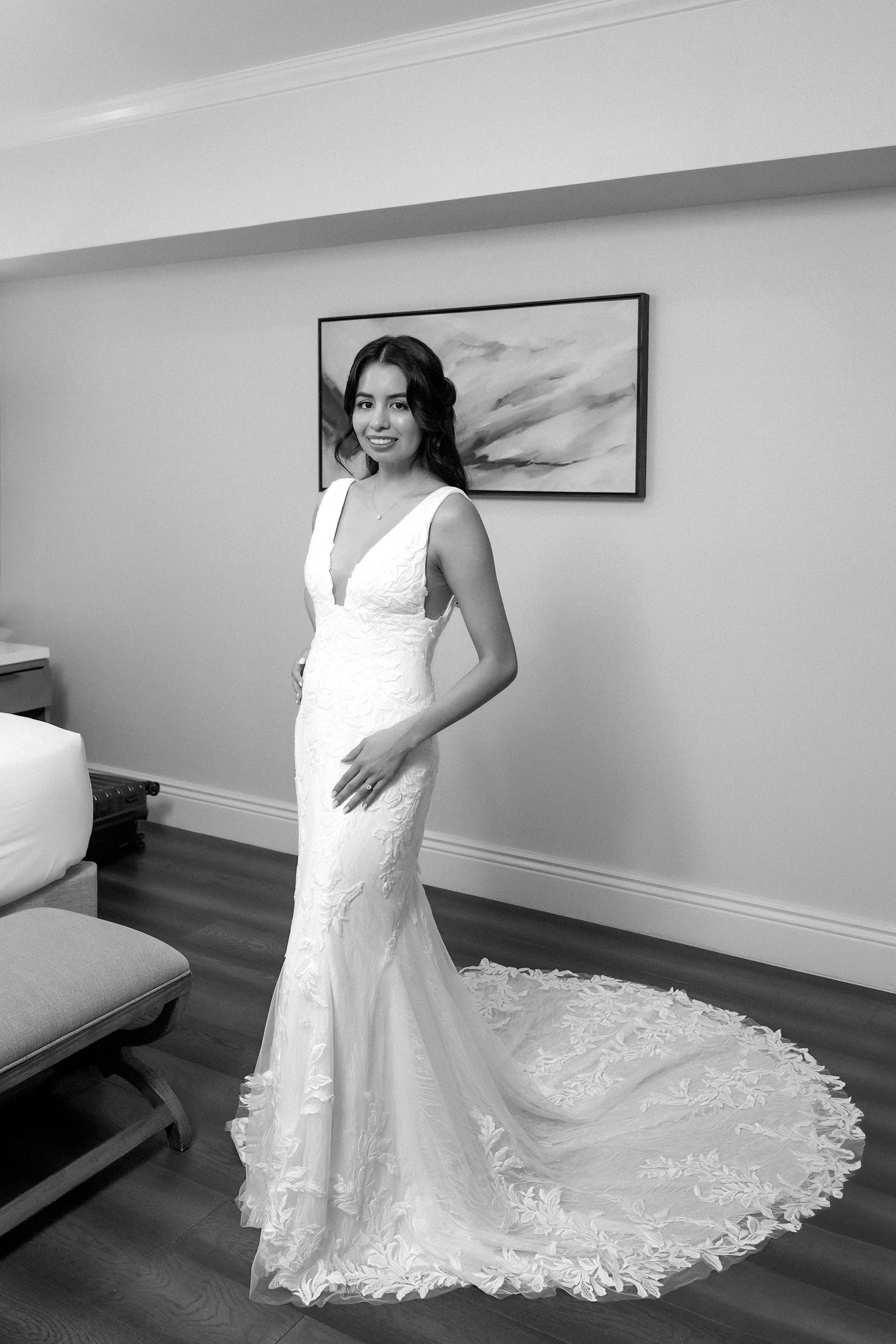A stunning black and white portrait highlighting a modern bride in her elegant wedding attire. She is wearing a fitted, mermaid-silhouette lace wedding gown featuring a dramatic, plunging v-neckline. The dress is adorned with intricate floral embroid