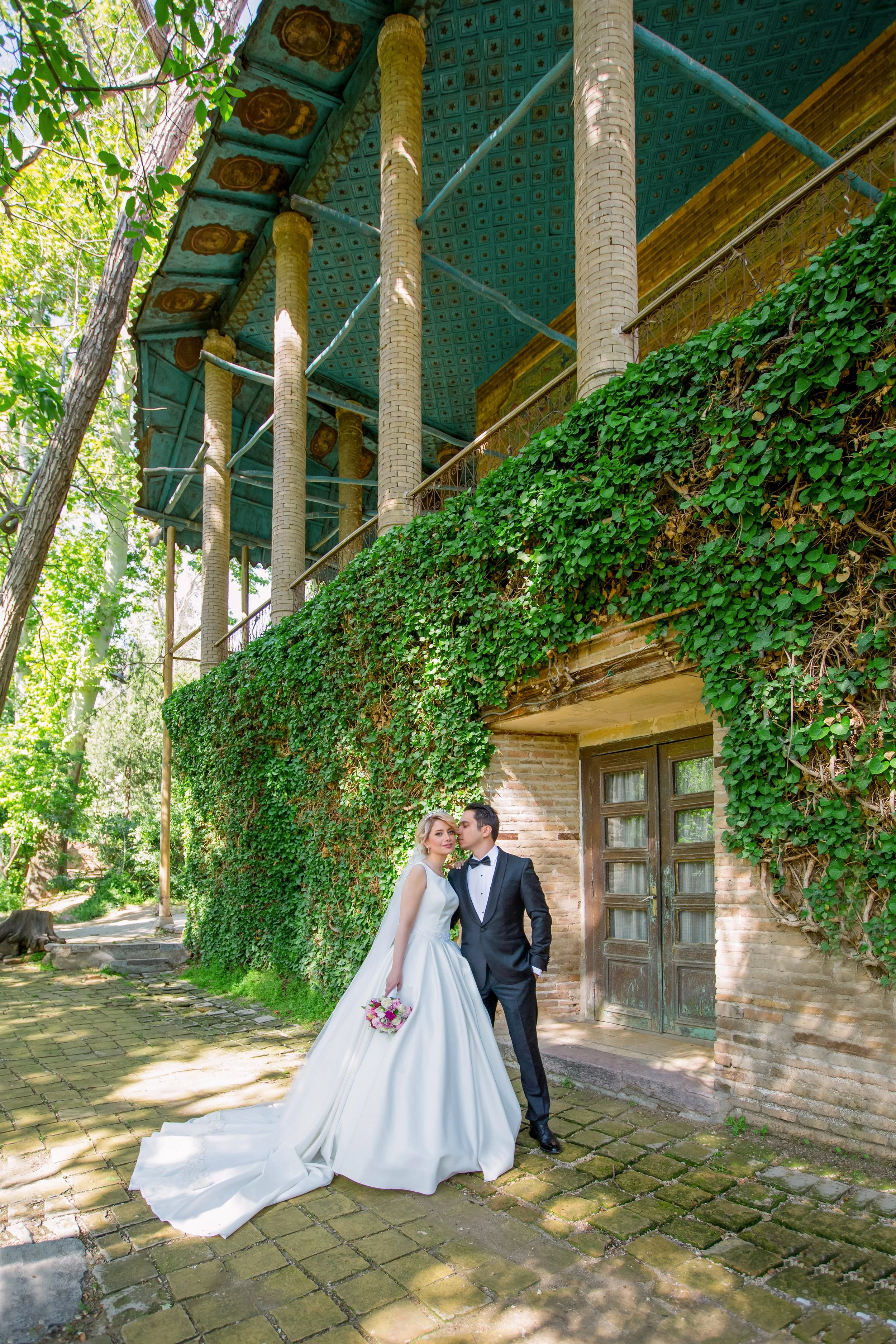 Bride and groom posing outdoors by an Ivy-covered brick building with an ornate blue tiled ceiling pavilion during wedding portraits.