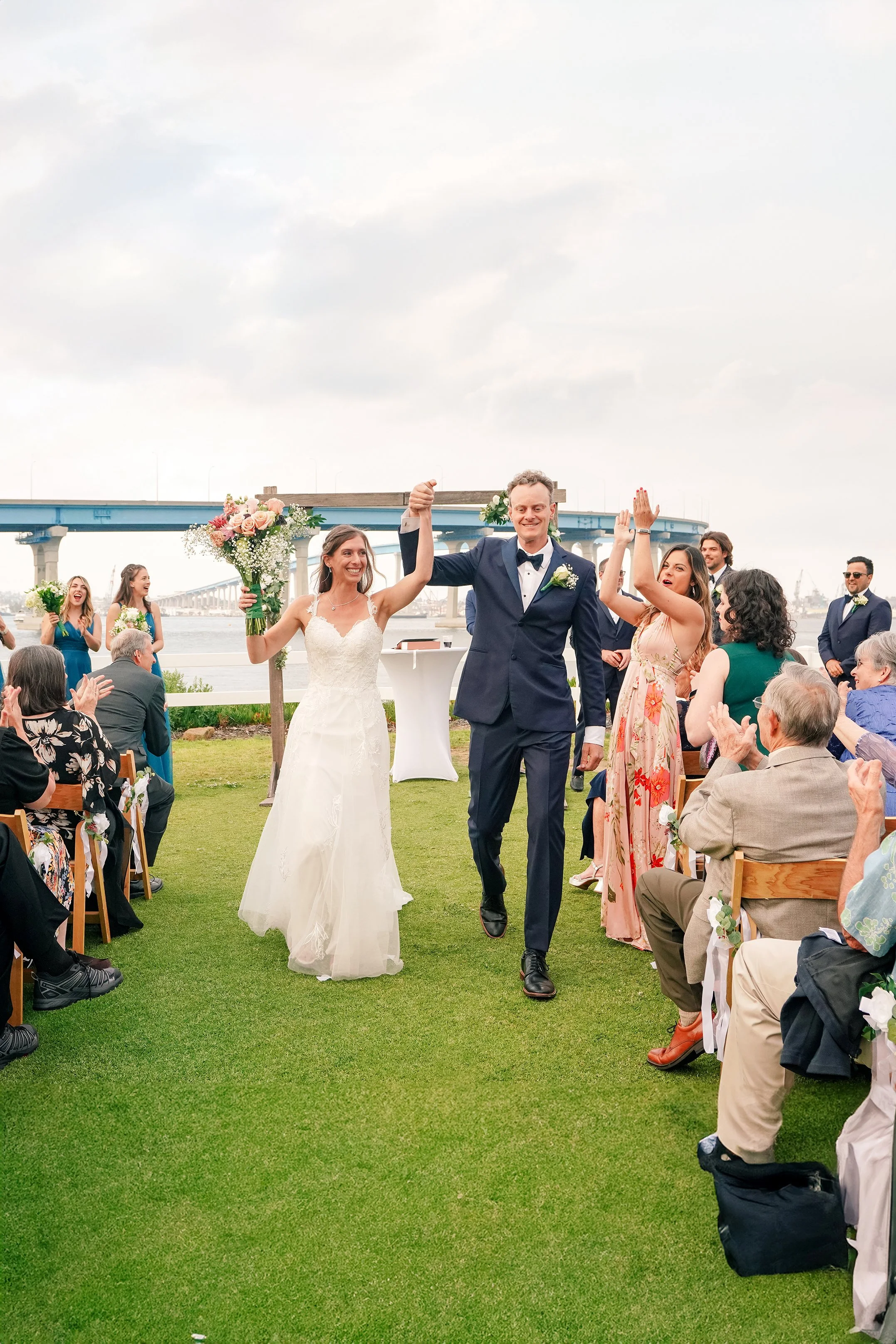 A full-length vertical shot of a newly married bride and groom walking up the aisle at an outdoor wedding. They are holding hands and lifting their arms in celebration while guests on both sides clap. The bride wears a white lace gown and holds her b