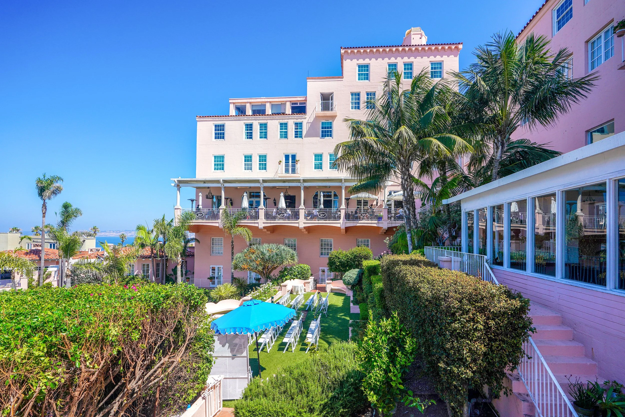 Wide view of the historic La Valencia Hotel exterior in La Jolla California showing the pink building, terrace, and an outdoor wedding ceremony setup on the garden lawn overlooking the Pacific Ocean.