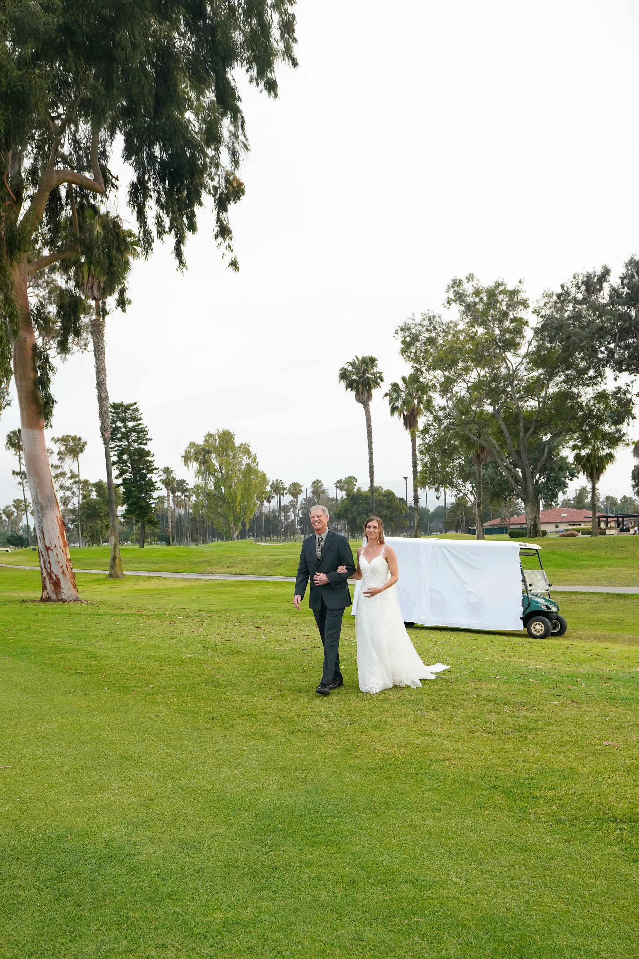 A bride and an older man, possibly her father, walking on a golf course with green grass and tall trees. The bride is wearing a white wedding dress, and the man is dressed in a suit. There is a golf cart and a covered vehicle in the background.
