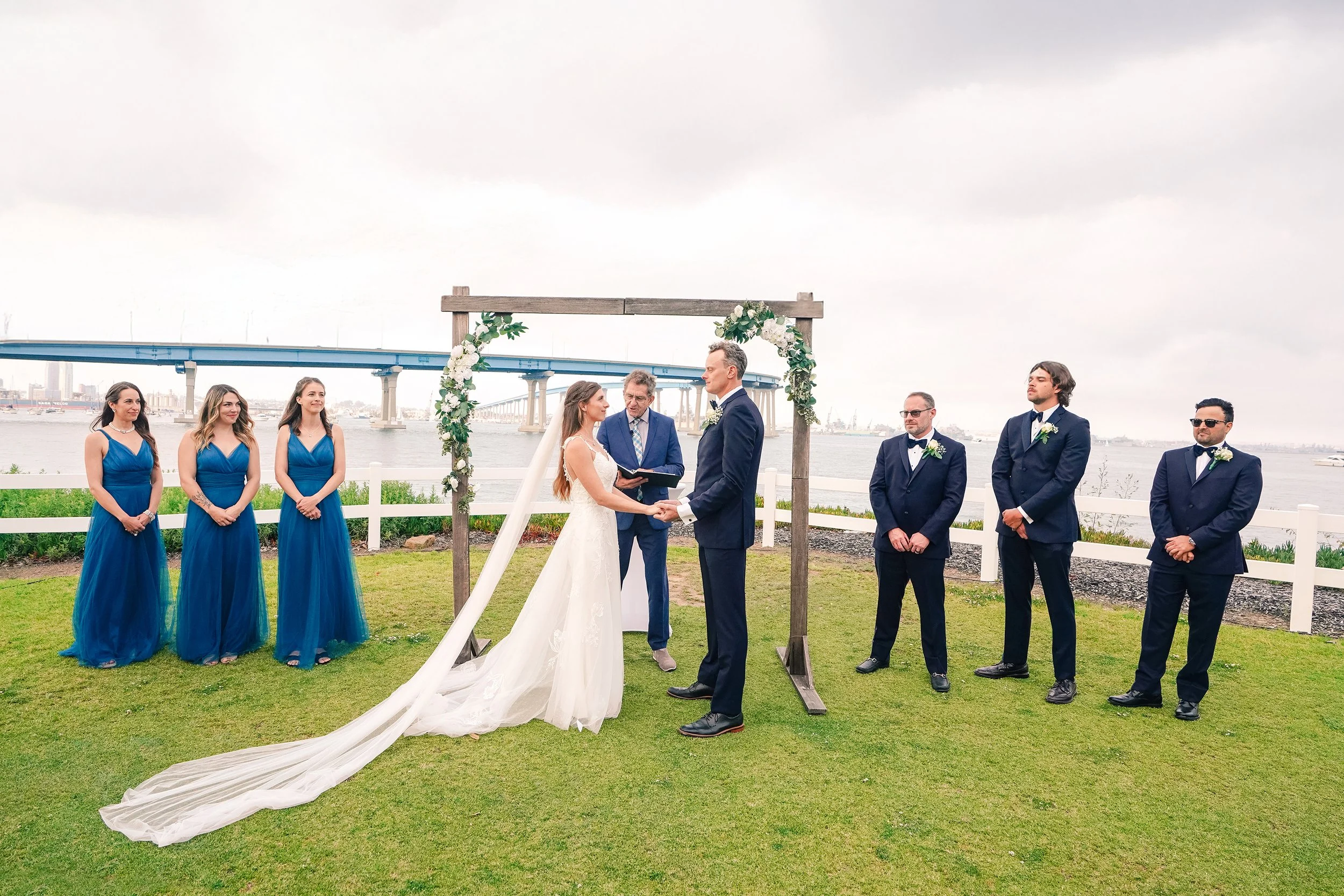 A couple gets married outdoors under a floral arch, with friends dressed in formal attire standing nearby, across a river with a bridge and city skyline in the background.