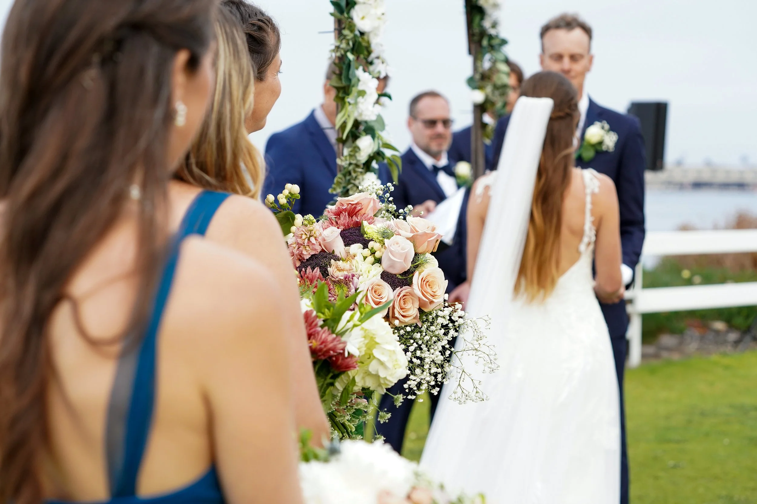 A wedding ceremony outdoors with a bride, groom, and guests. The bride is holding a bouquet of pink, white, and purple flowers. The groom and guests are dressed in formal attire, with some men wearing suits and women wearing dresses. The ceremony is 