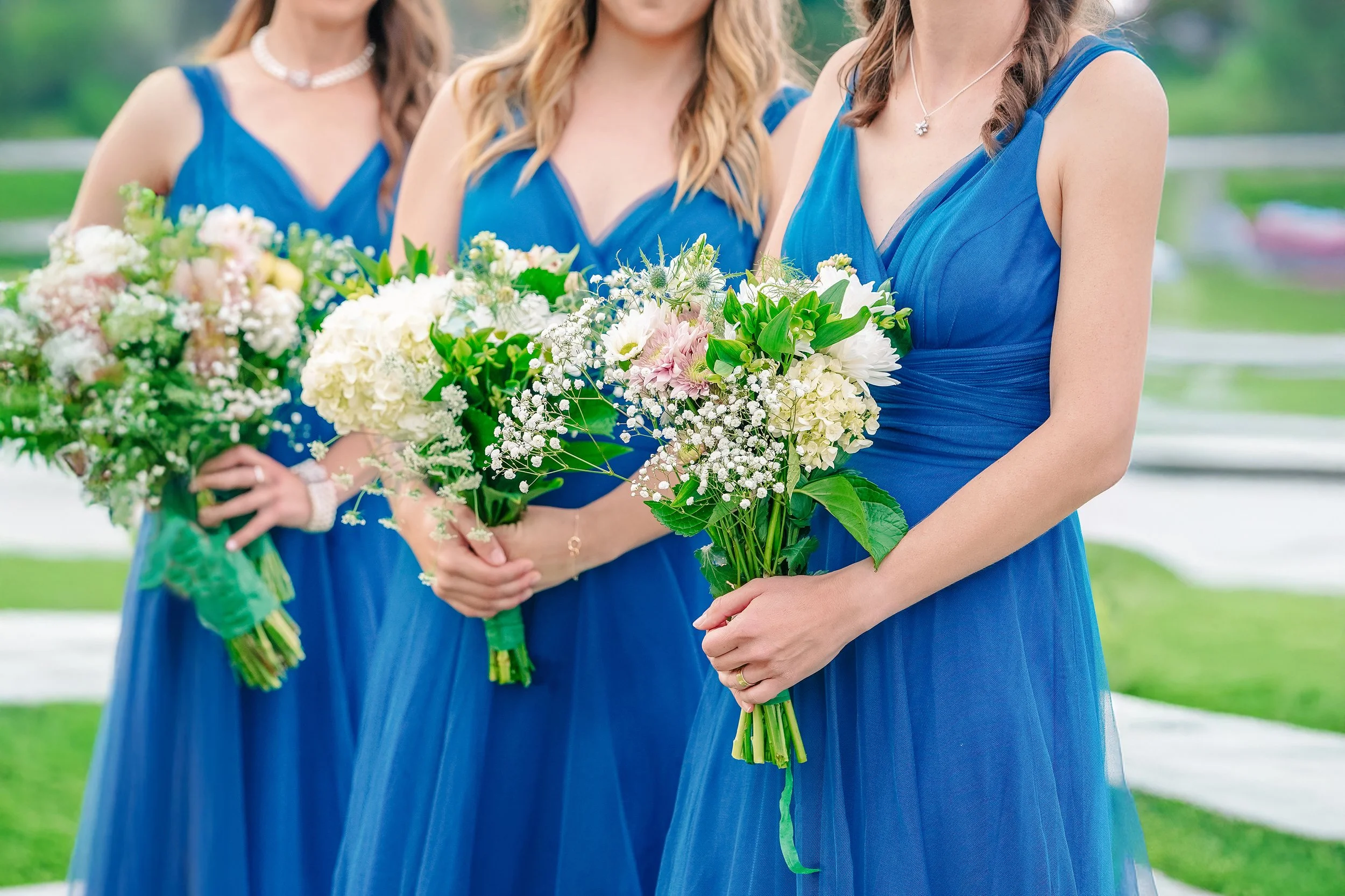 Women in blue dresses holding bouquets of white, pink, and green flowers at an outdoor event.
