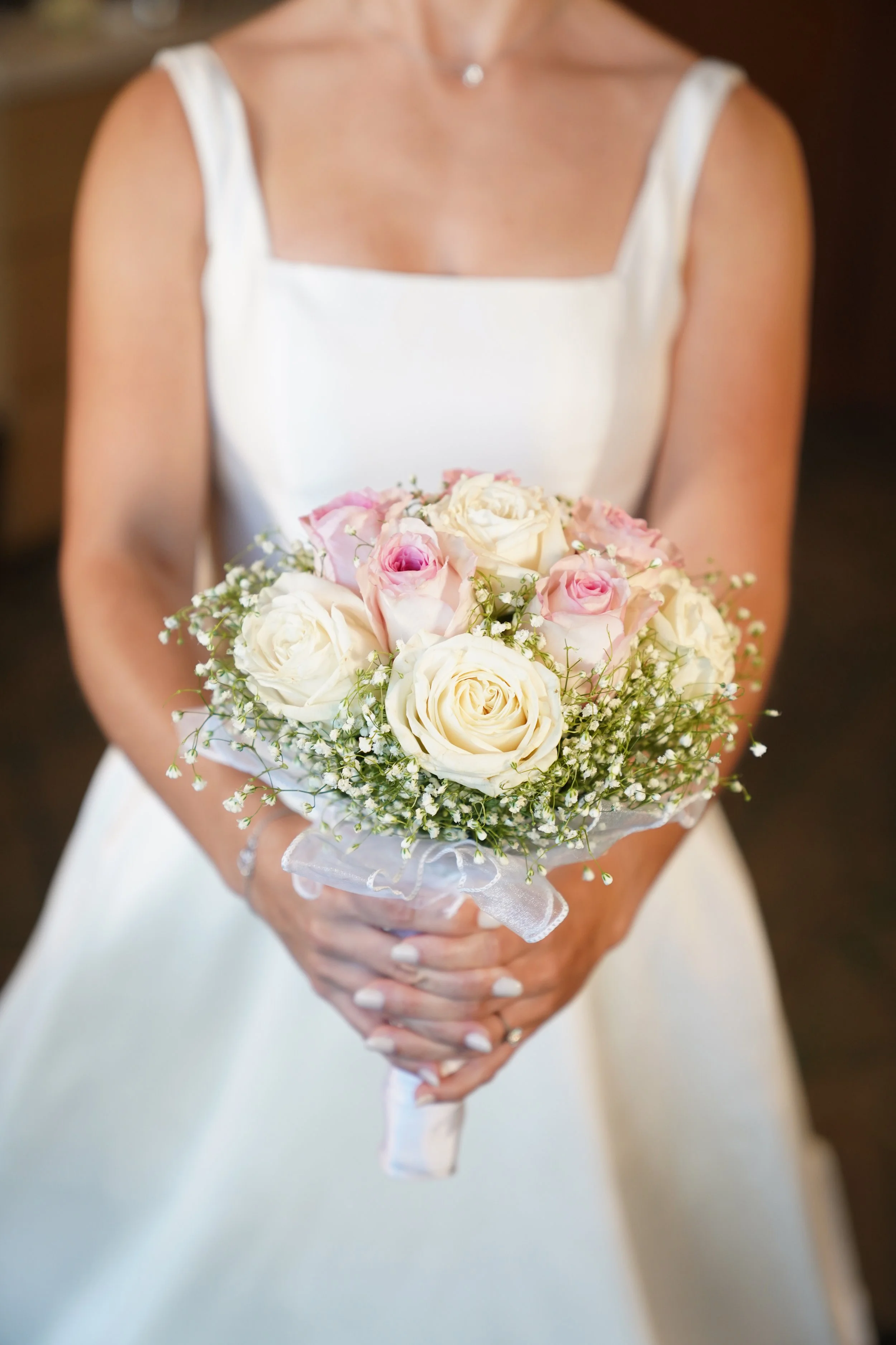 Close-up of a bride holding a round wedding bouquet made of light pink and white roses wrapped in baby's breath.