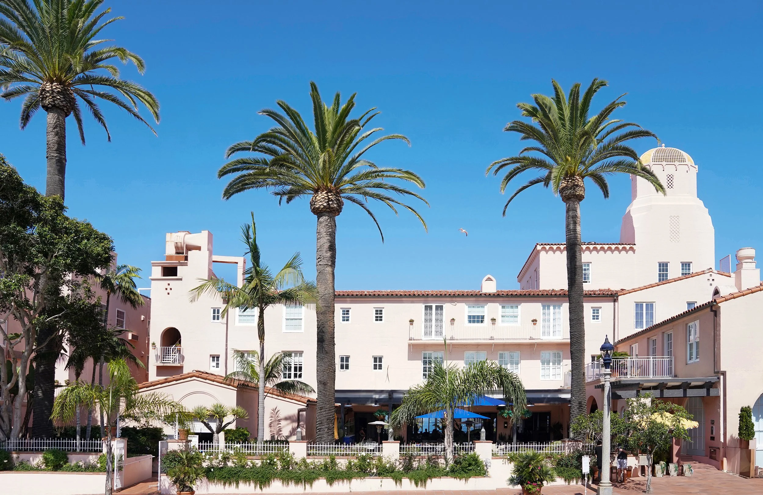 Wide exterior view of the pink La Valencia Hotel resort with tall palm trees, Spanish revival architecture, and a clear blue sky in La Jolla, San Diego.