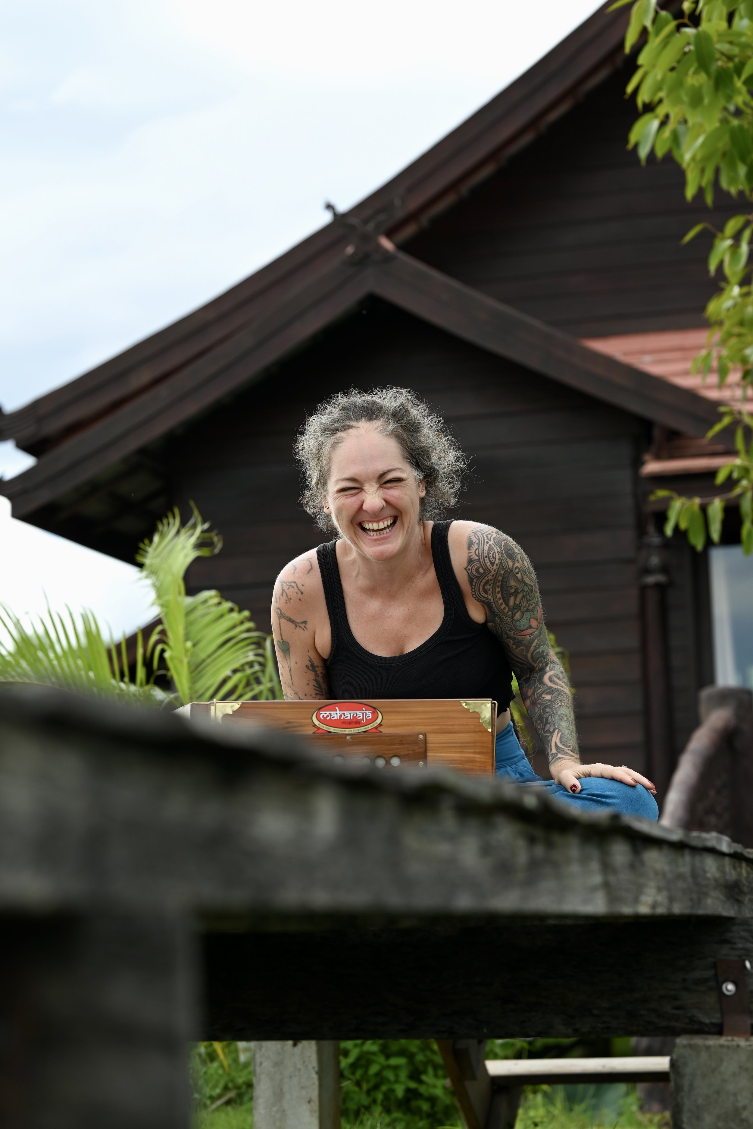 A woman with curly gray hair and tattoos, wearing a black tank top, is smiling joyfully while playing a wooden arched kalimba outdoors, with a dark wooden house and green foliage in the background.
