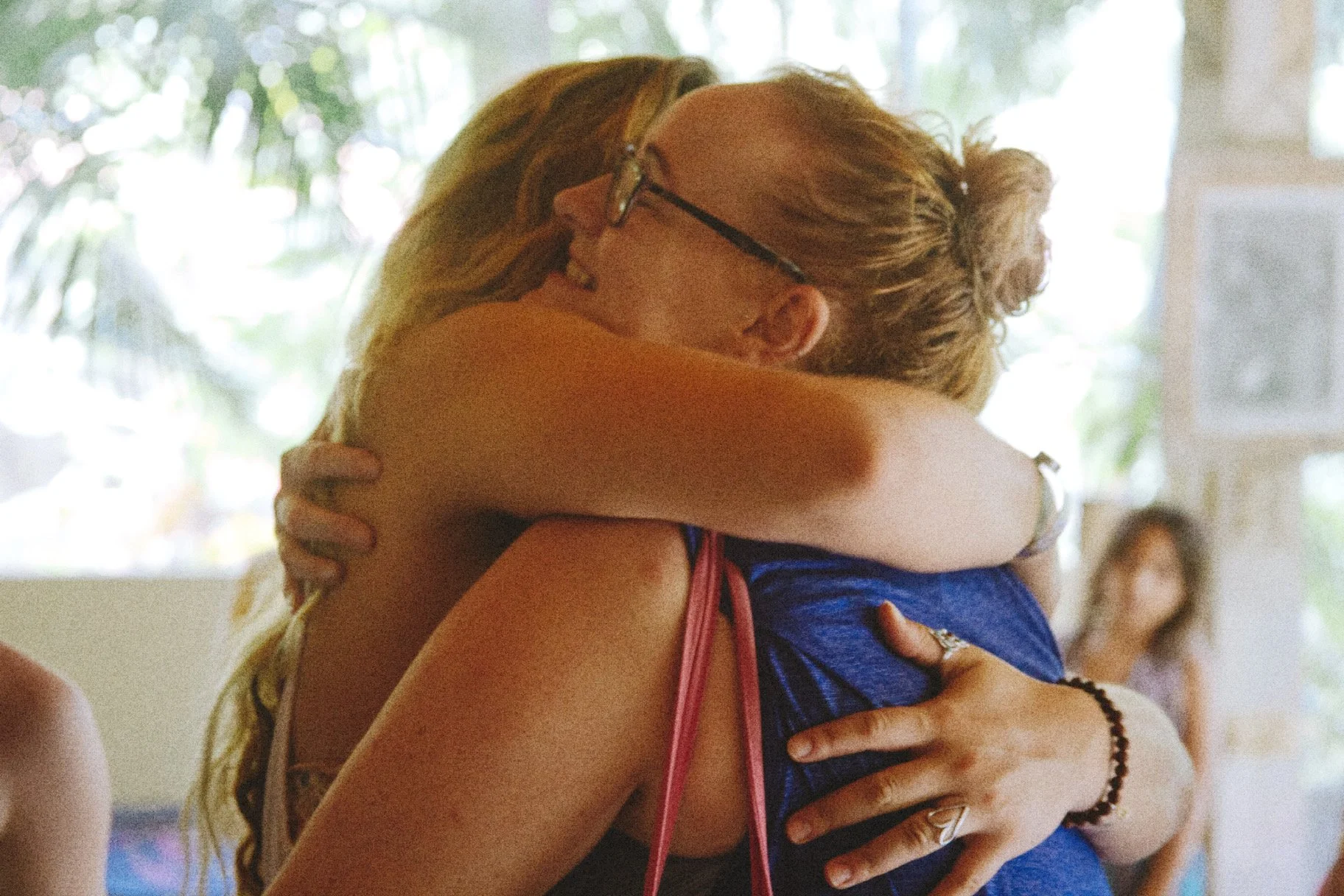 Two women hugging each other tightly in a loving embrace indoors, with sunlight coming through windows in the background.