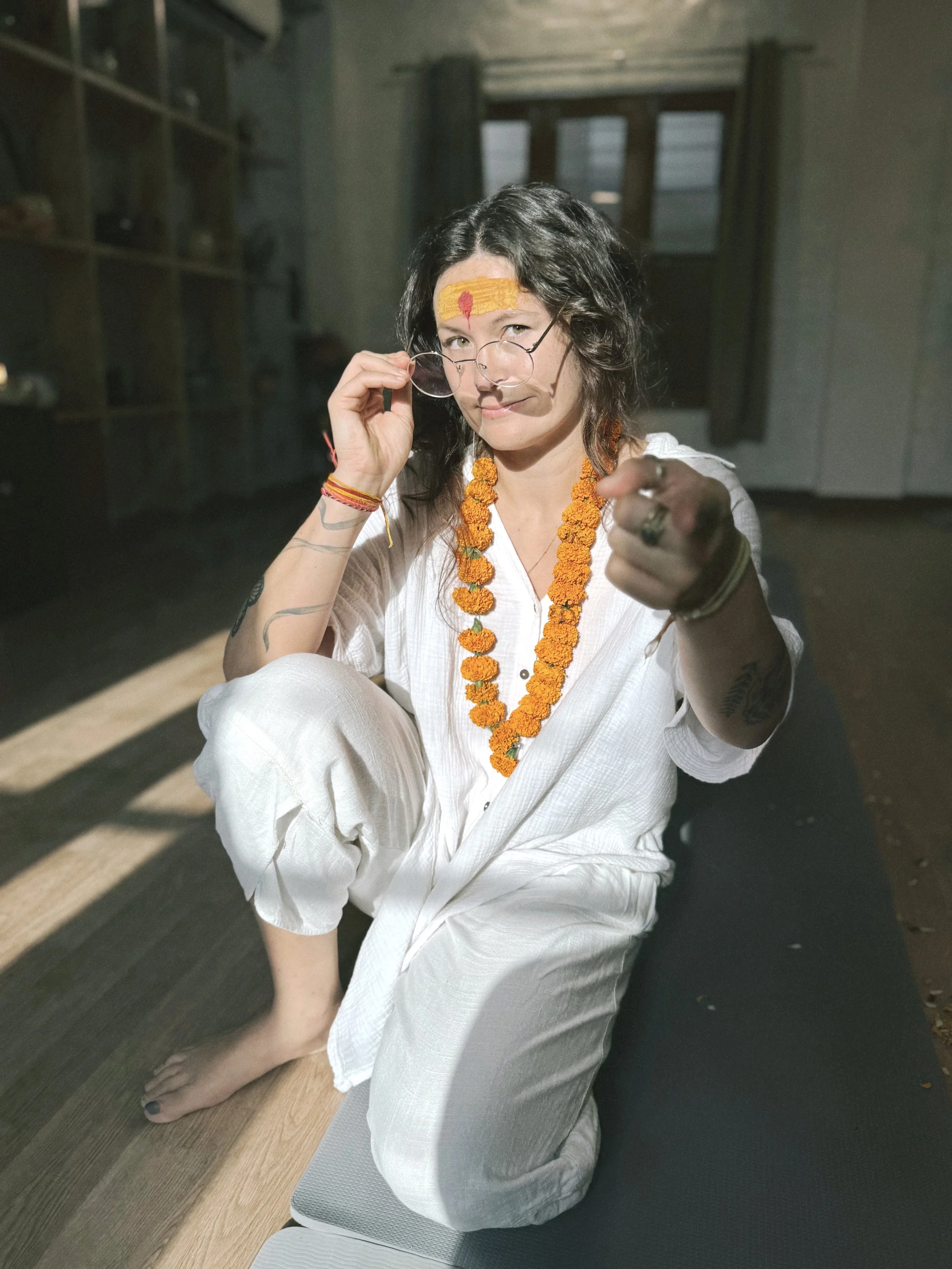 Woman with tattoos and glasses, sitting on a yoga mat, wearing white clothing, with marigold garlands and a painted forehead, inside a room.