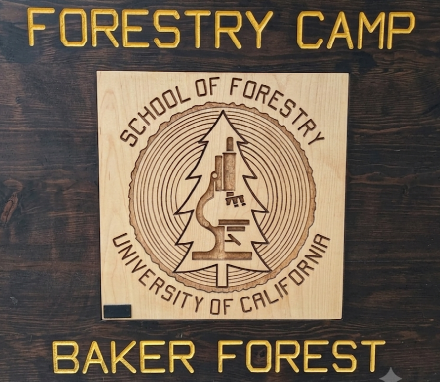 Wooden plaque with the logo of the School of Forestry at the University of California, featuring a tree silhouette and a microscope, and surrounded by concentric circles. Golden text at the top reads "Forestry Camp" and at the bottom "Baker Forest" in gold lettering.