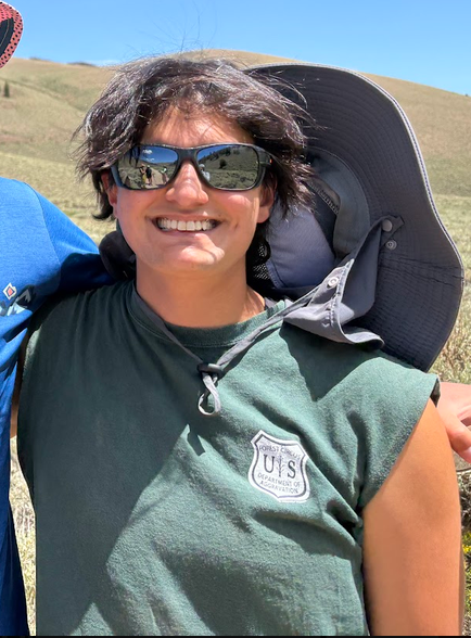 Nonbinary person with short curly hair wearing sunglasses, a green sleeveless shirt with a U.S. Forest Service logo, smiling outdoors in a grassy, hilly landscape.