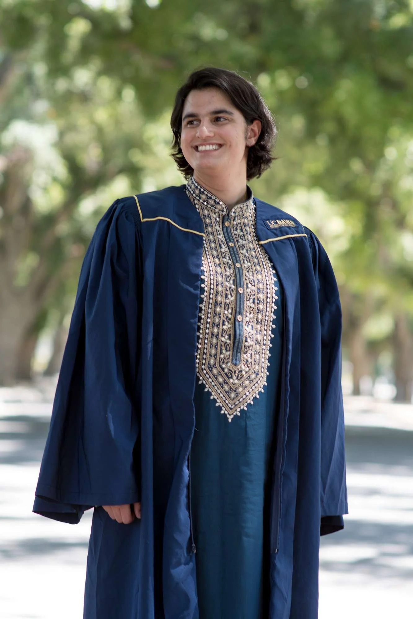 Young person in a blue UC Davis graduation gown and traditional indian kurta, smiling, outdoors with green trees in the background.