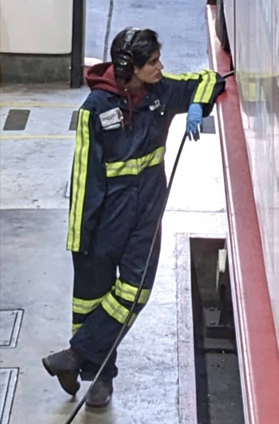 A person with short dark hair stands in a repair shop, leaning against a bus lift, wearing blue coveralls with reflective stripes, blue nitrile gloves, and ear protection.