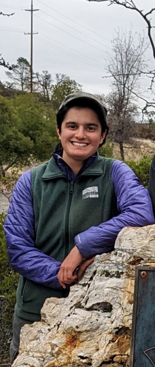 Young person smiling outdoors, wearing a dark green vest over a purple jacket, with trees and power lines in the background.