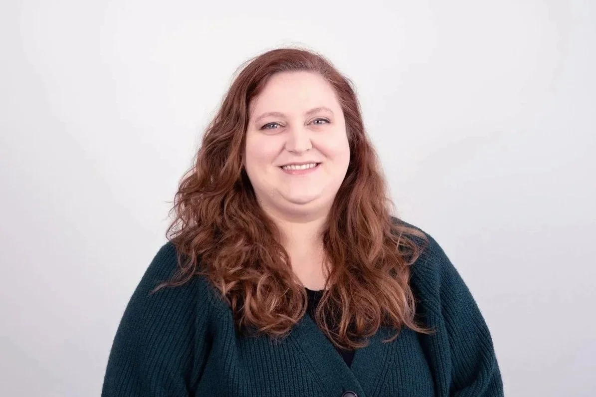 A woman with long curly red hair smiling at the camera, wearing a dark cardigan against a plain white background.