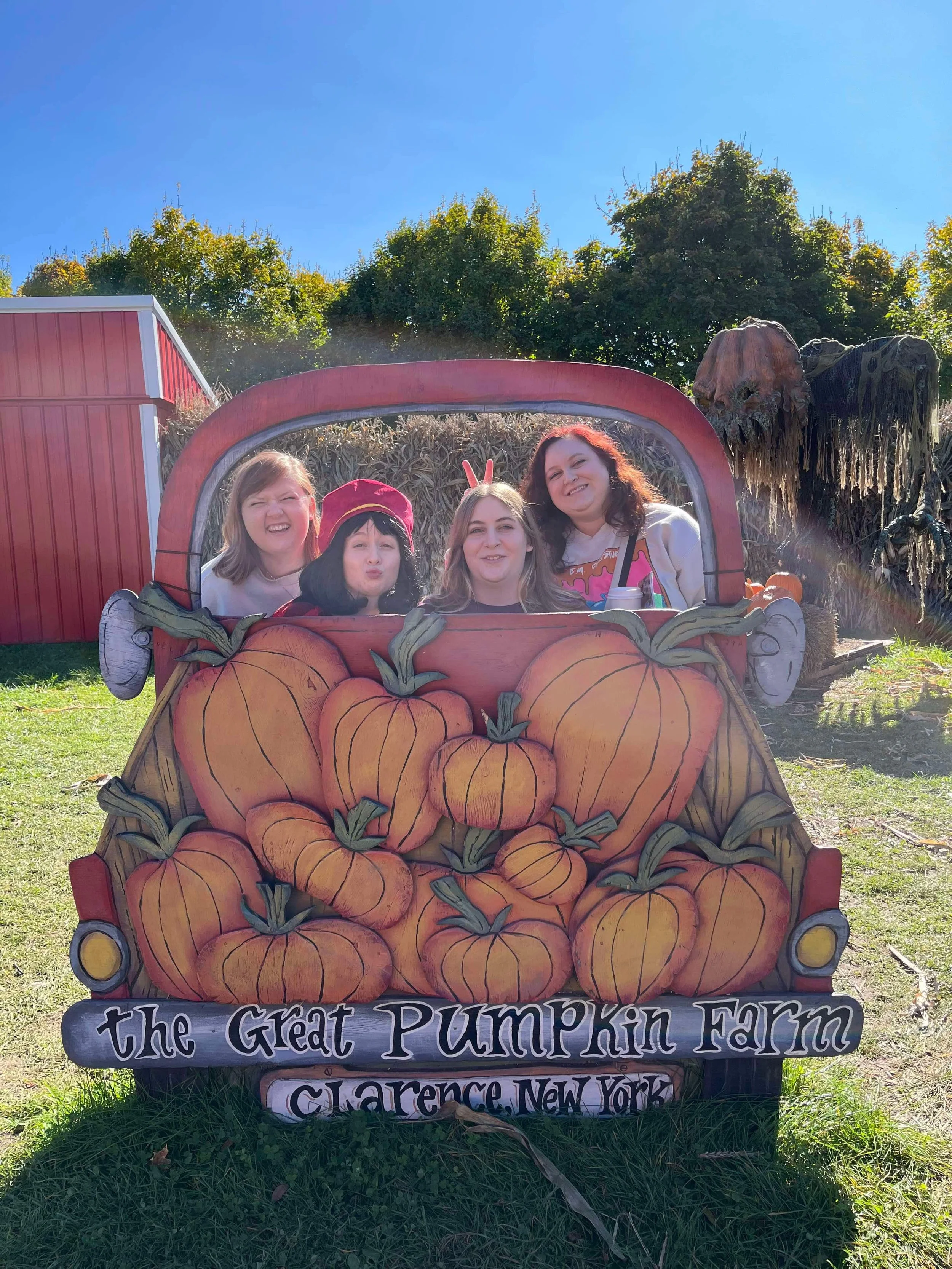 Sign for the Great Pumpkin Farm in Clarence, New York, featuring large decorated pumpkins and cartoonish pumpkin graphic, with a display of four smiling women posing behind it, autumn decorations in the background, and a bright blue sky.