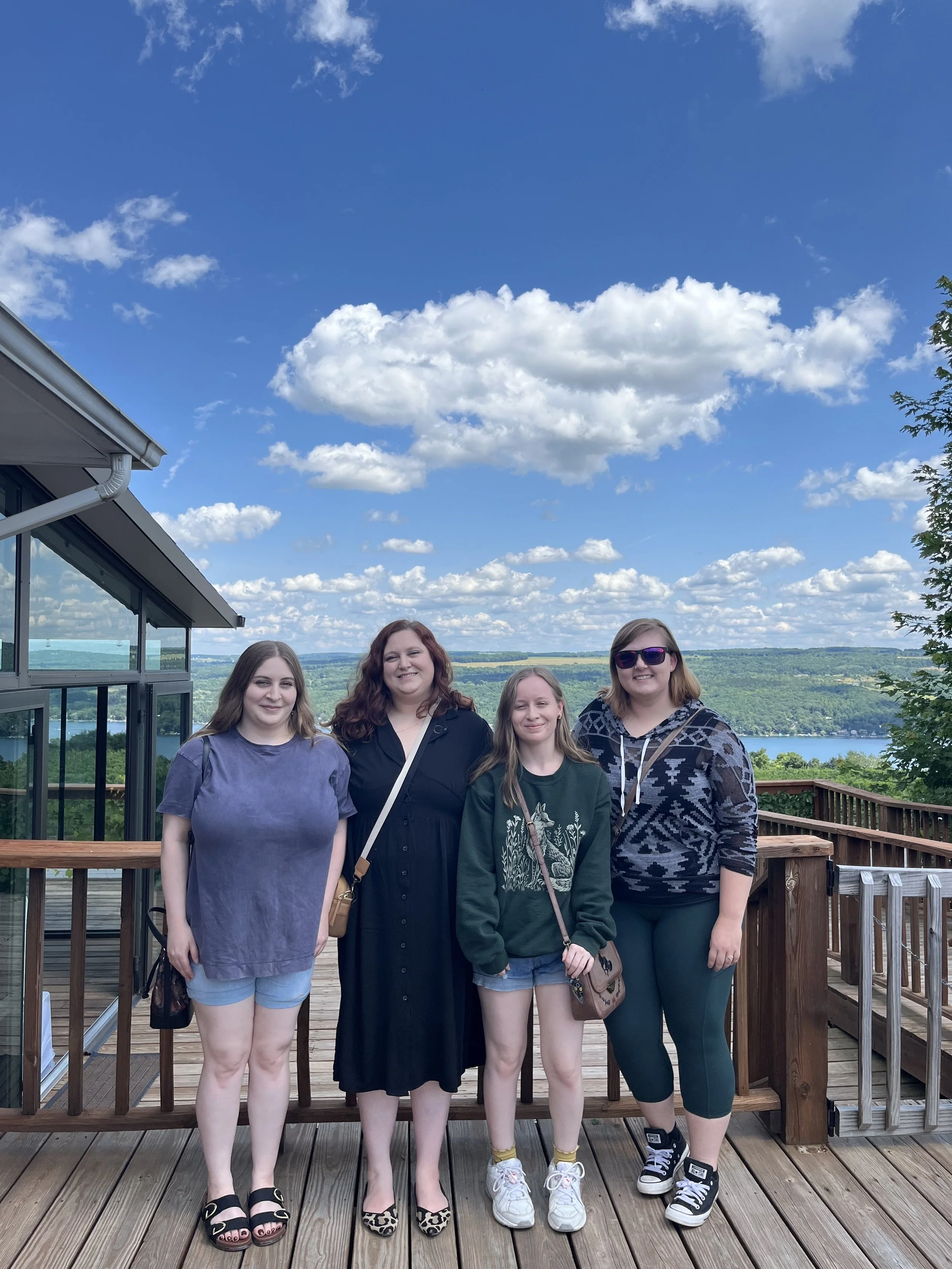 Four women standing on a wooden deck outdoors, smiling, with a scenic lake and green hills in the background under a bright blue sky with scattered white clouds.