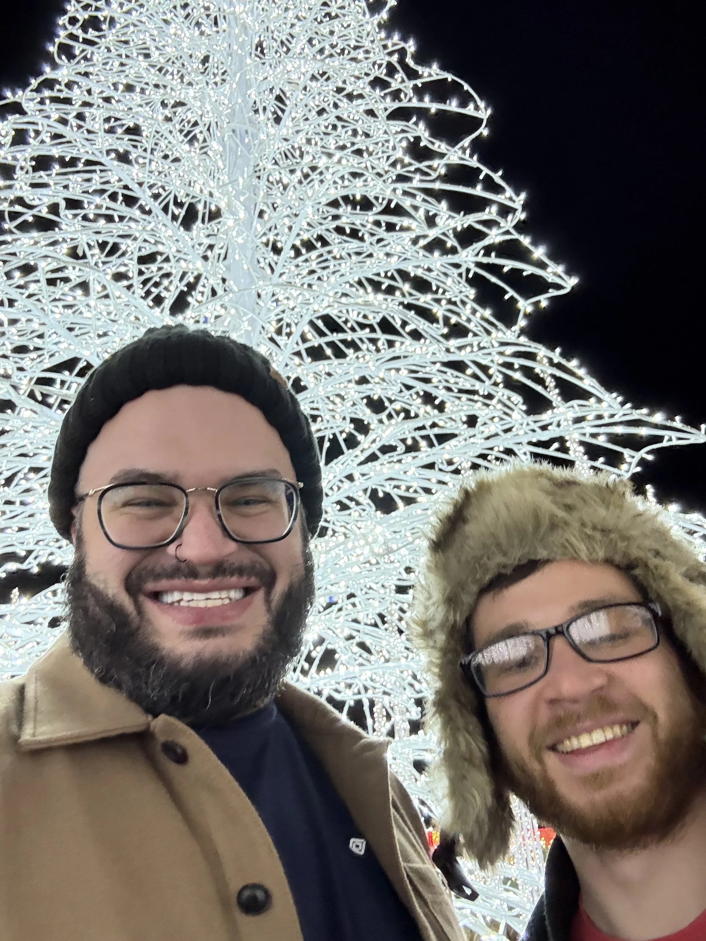 Two smiling men taking a selfie in front of a large illuminated decorative tree at night.