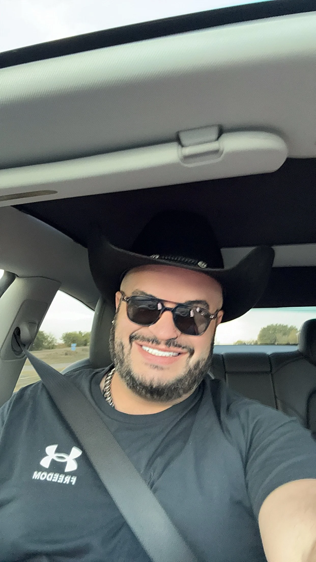 A man with a beard and sunglasses is smiling and taking a selfie in the front seat of a car. He is wearing a black Under Armour t-shirt and a chain necklace, with a black cowboy hat. The background shows a clear sky and open land.