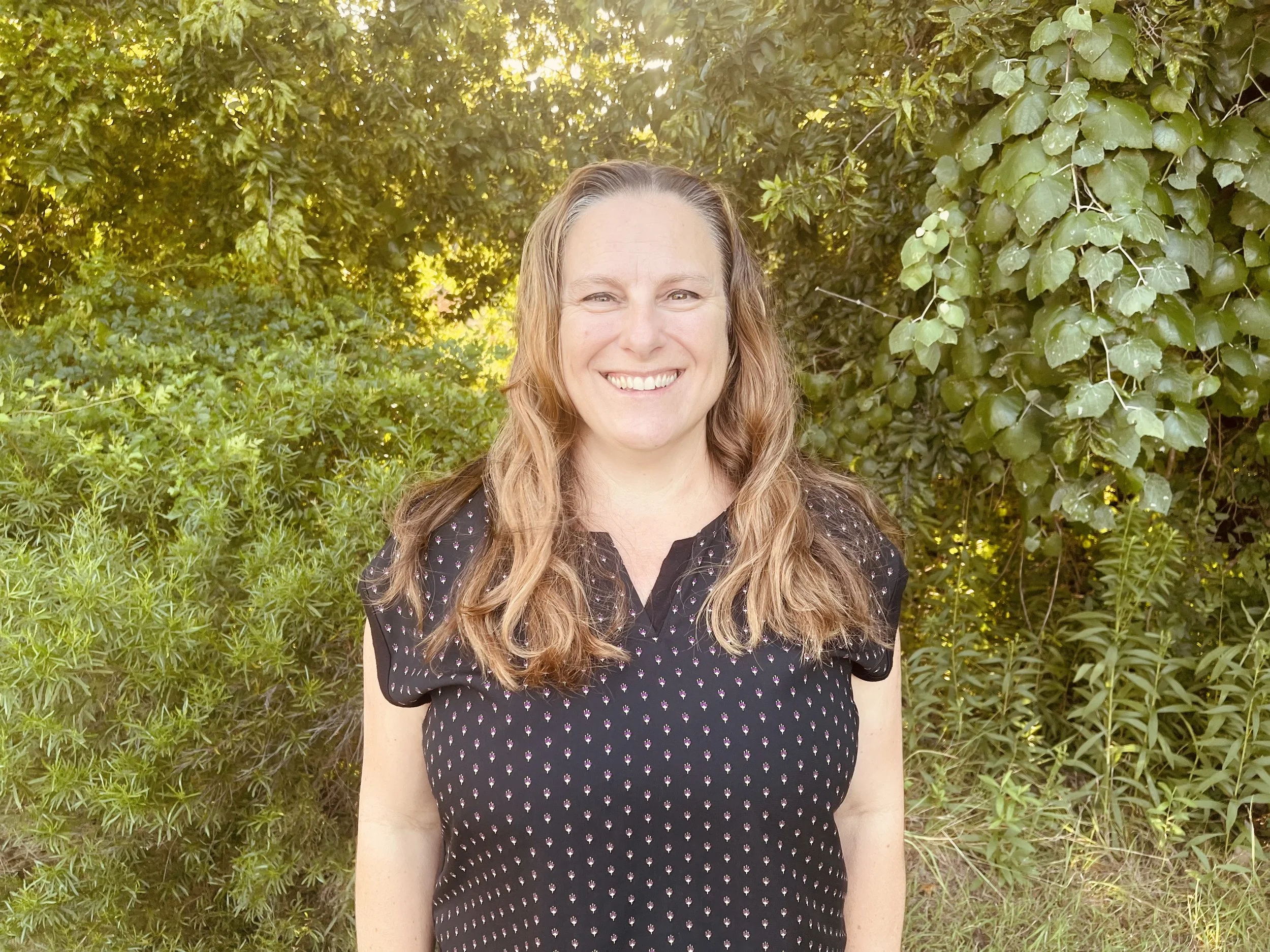 A woman with long, wavy hair smiling outdoors in front of lush green foliage, sunlight filtering through the trees.