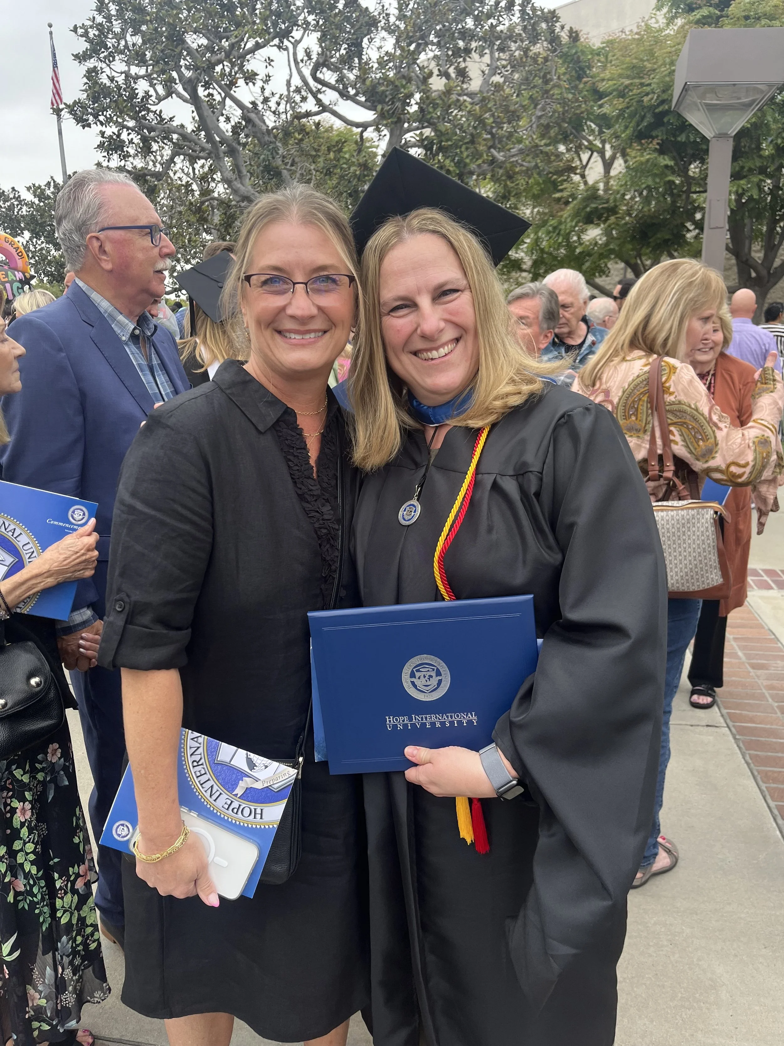 Two women smiling at a graduation ceremony, one in a black academic gown and the other in a dark jacket, holding a blue diploma folder from Hope International University, surrounded by other graduates and attendees.