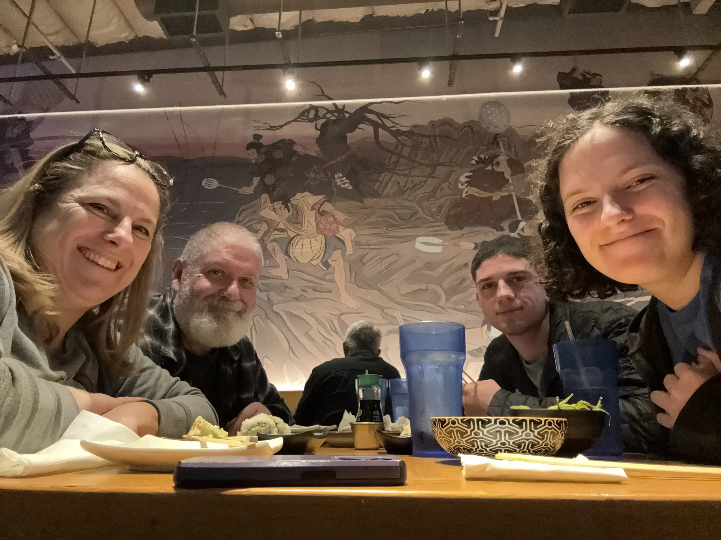A group of six people sitting around a table in a restaurant, smiling at the camera. The background features a mural with stylized tree and human figures.