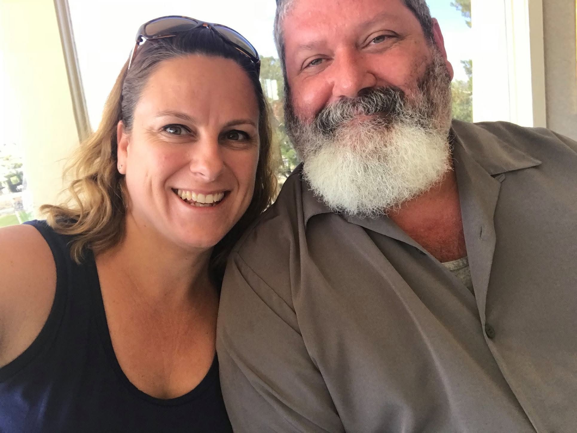 Close-up photo of a woman with sunglasses on her head and a man with a full white beard, both smiling, taking a selfie indoors with sunlight and trees visible through the window behind them.