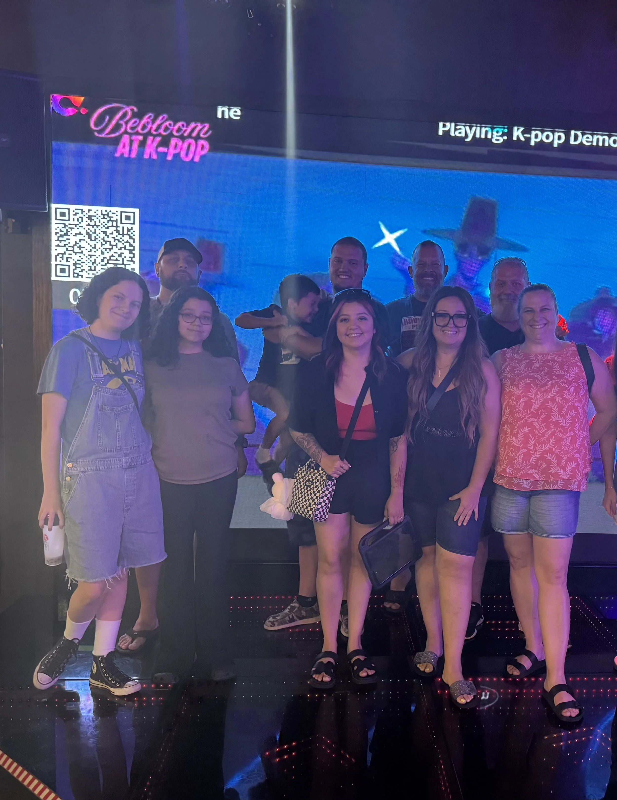 Group of nine people standing in front of a digital screen displaying a space-themed image and text about a K-pop demonstration. Some are smiling, with one person holding a drink, and another holding a small purse.