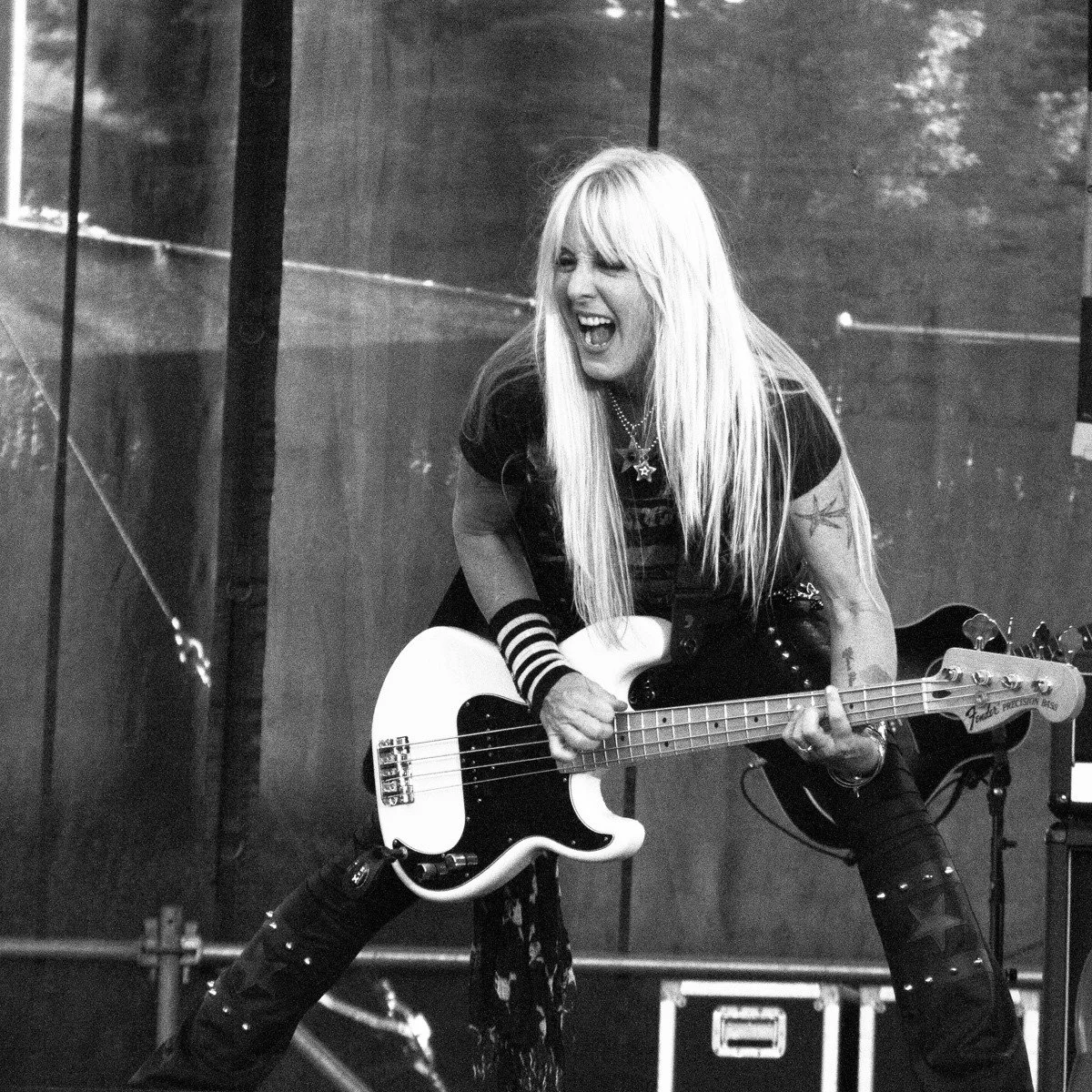A woman with long blonde hair, wearing dark clothing with star designs and striped wristbands, playing an electric bass guitar and singing passionately on stage.