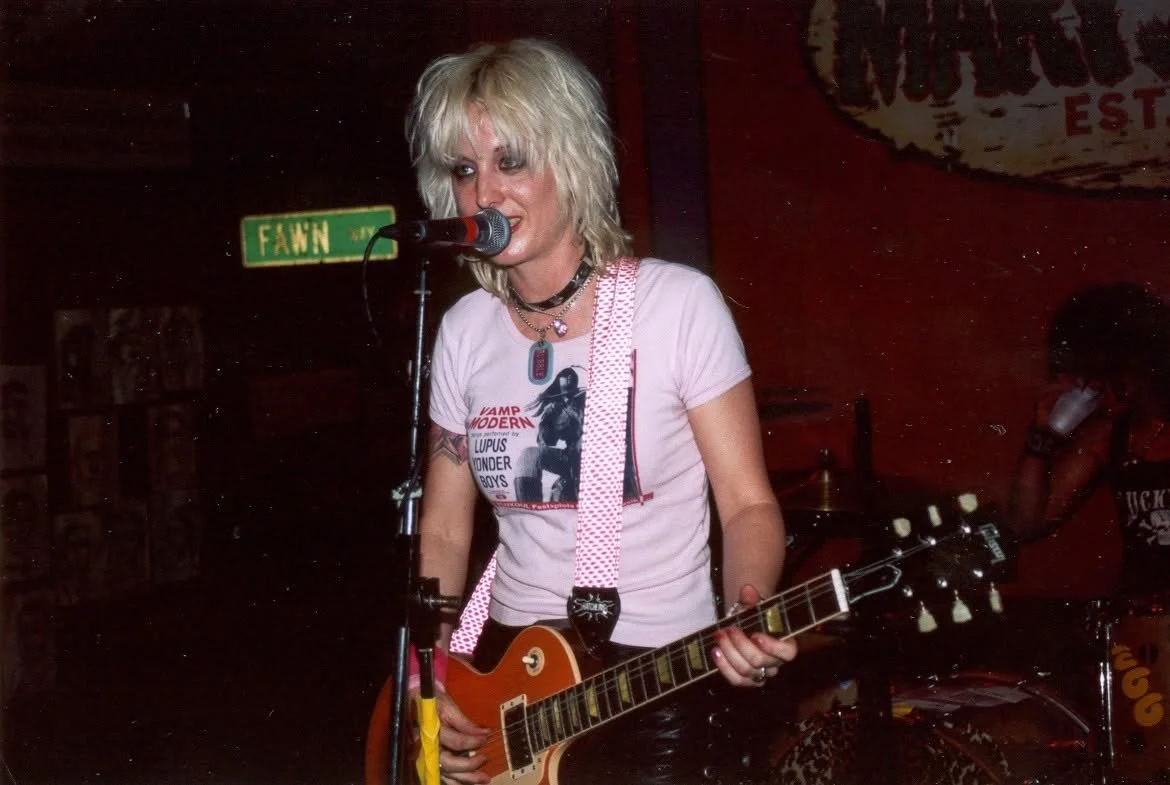 A woman with blonde, messy hair singing into a microphone while playing an electric guitar on stage. She is wearing a white t-shirt with a graphic print, layered necklaces, and a polka dot guitar strap, with a red wall and a band member in the backgr