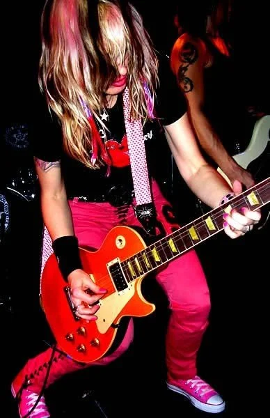 A person with long, blonde hair with pink highlights playing a red electric guitar. They are wearing a black T-shirt, pink pants, and pink sneakers, with a strap over their shoulder and a black wristband on one arm.