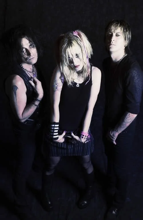 Three people with punk or goth style clothing and makeup standing against a dark background.